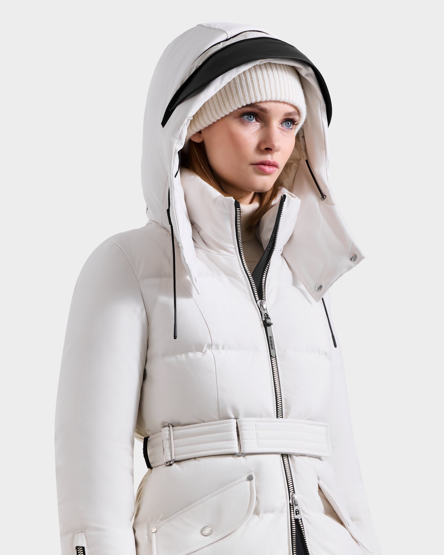 A woman wearing the Rudsak CALI H - 8125614 Vanilla women’s long down parka with a belted waist and removable hood, paired with a white knit beanie, looks slightly to the side against a plain light background.