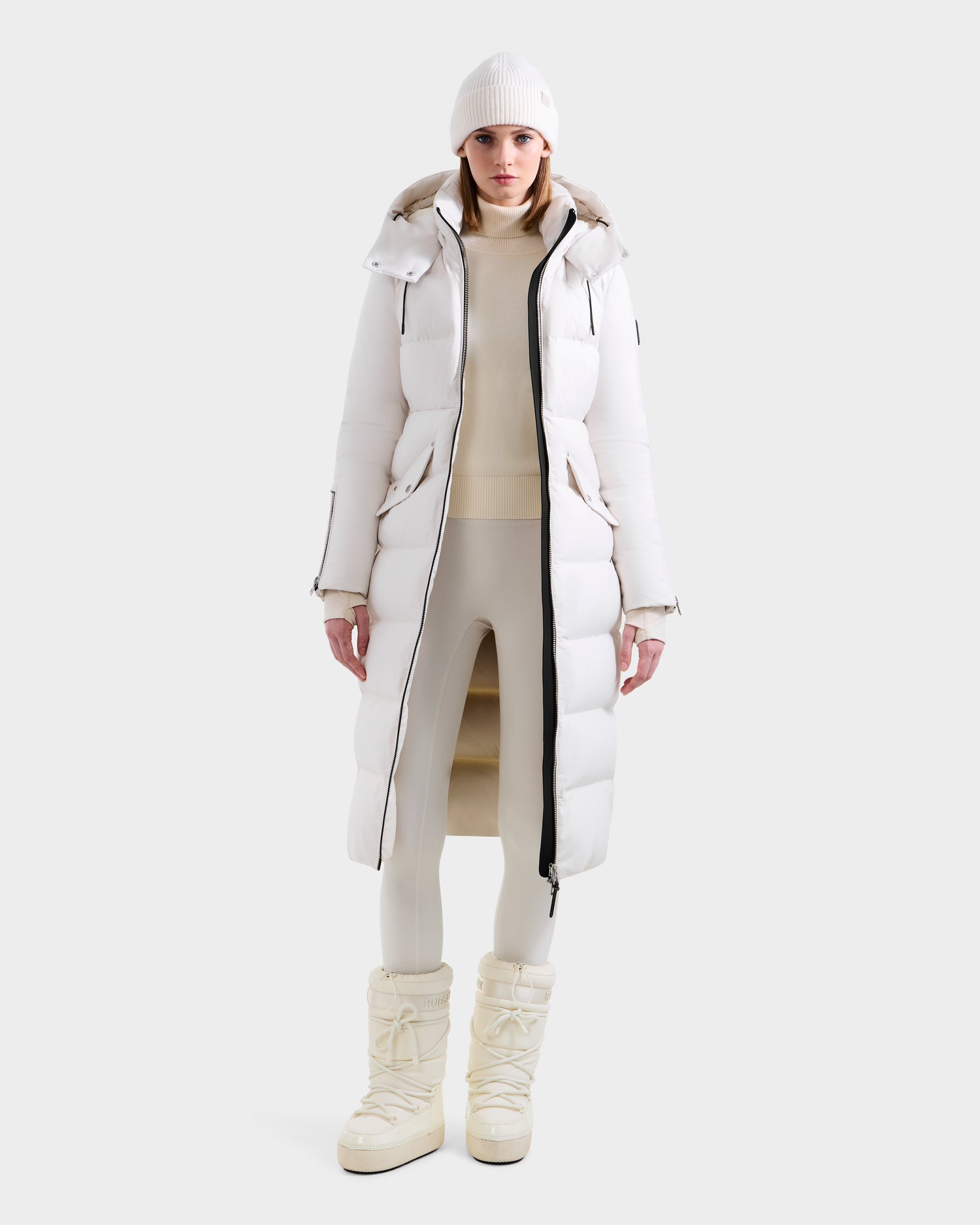 A person stands facing forward against a light background wearing the Rudsak CALI H - 8125614 Vanilla women’s long down parka with high-loft down and removable hood, styled with a white knit beanie, light turtleneck, fitted pants, and white winter boots.