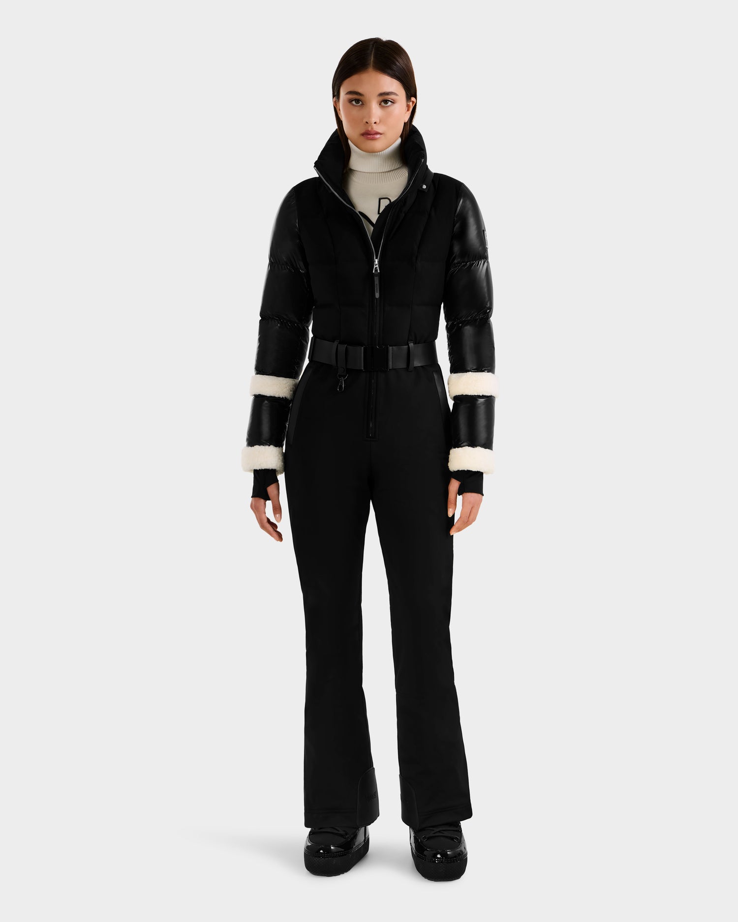A woman faces forward in the Rudsak CARMEN - 8125708 Black fitted ski suit, featuring puffer sleeves, white shearling arm trim, a white turtleneck, and black boots—crafted for style and safety with integrated RECCO technology.