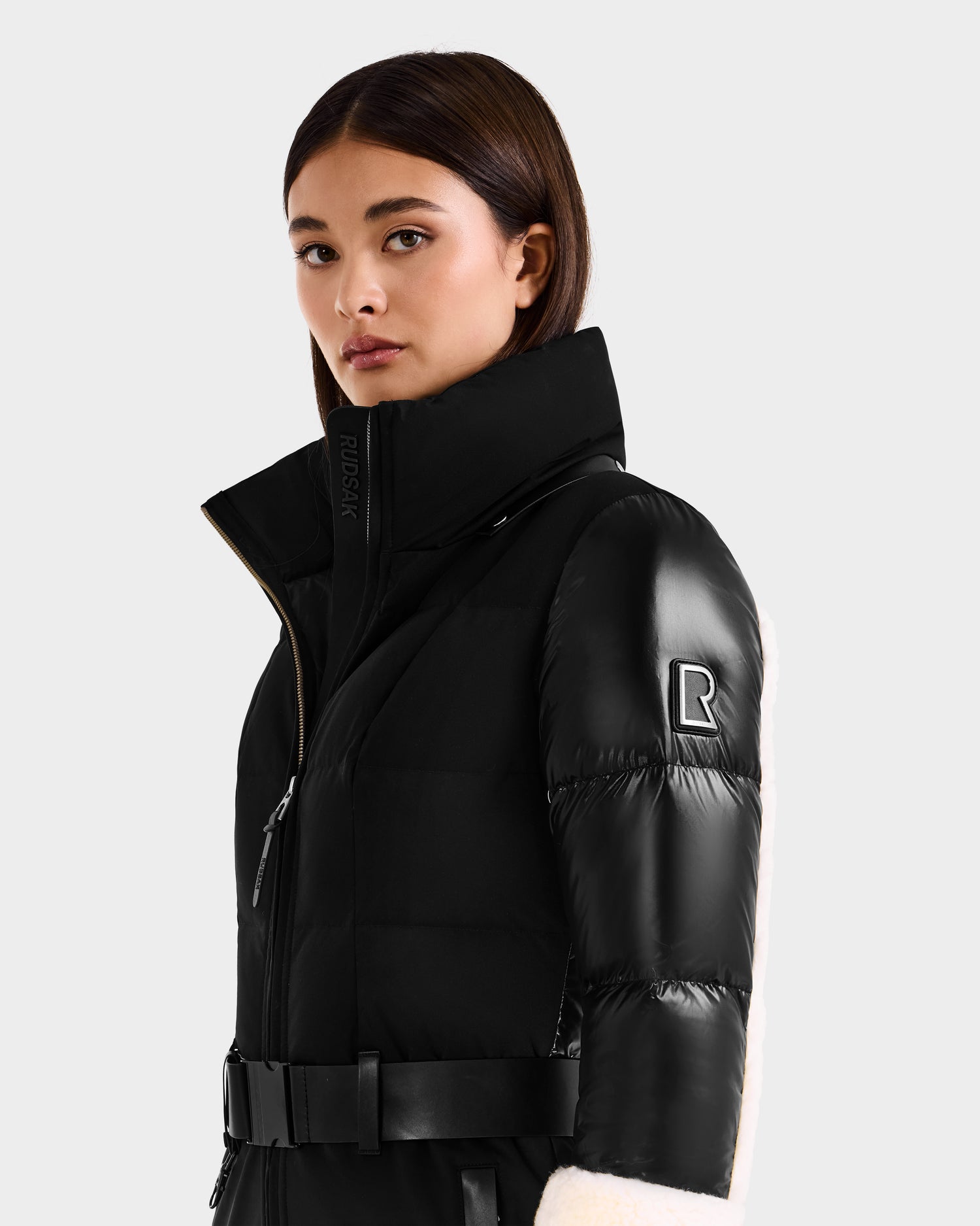 A woman wears the Rudsak CARMEN - 8125708 black puffer jacket with a belt, high collar, and sleeve logo, standing against a plain light gray background.