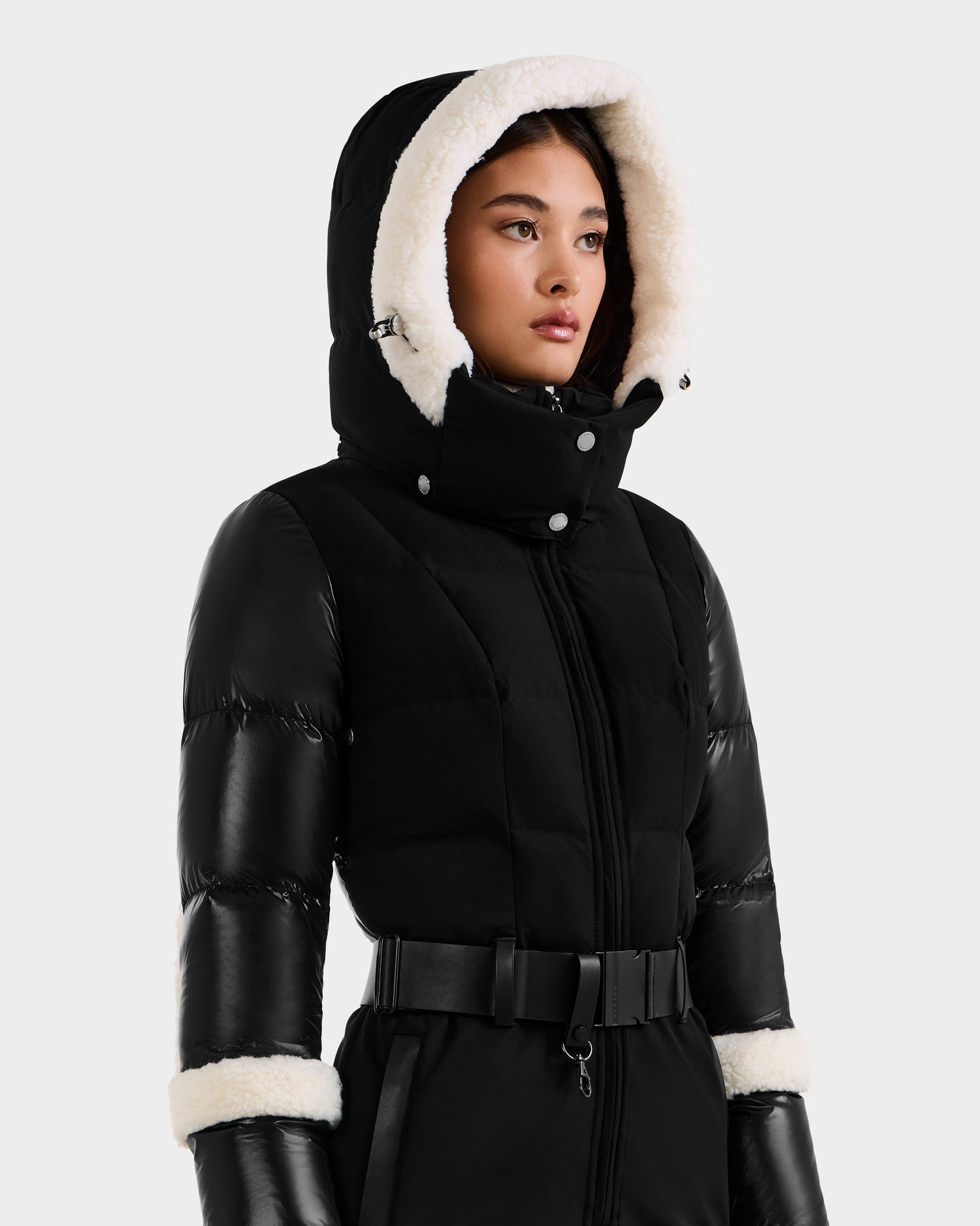 A woman wearing the Rudsak CARMEN - 8125708 Black puffer jacket with a belted waist and white fur-lined hood and cuffs, looking slightly to the side against a plain light background.