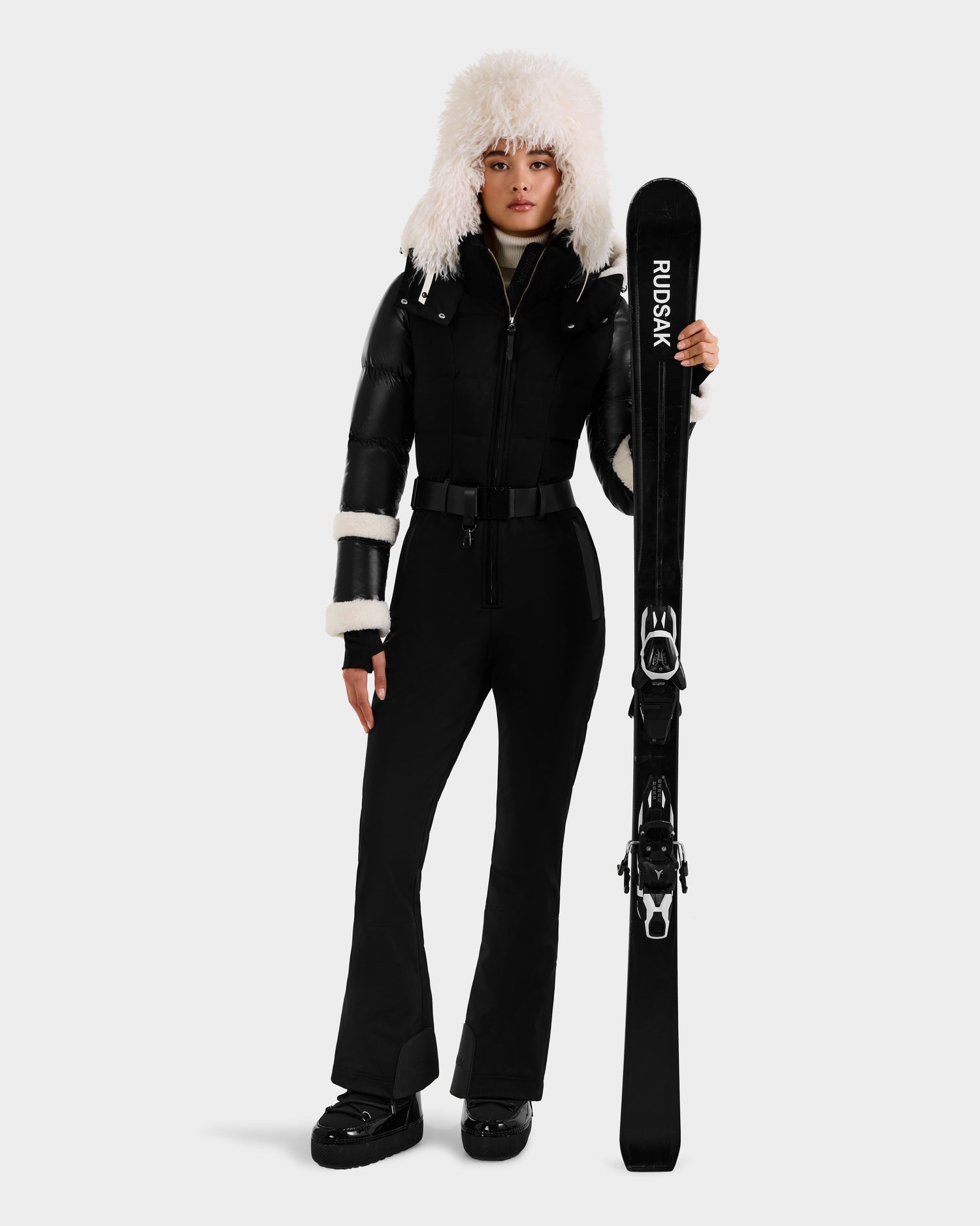 Dressed in the Rudsak CARMEN - 8125708 Black one-piece ski suit, black boots, and a fluffy white hat, a person stands holding black Rudsak skis. The plain light gray background highlights their sleek gear and integrated RECCO rescue technology.