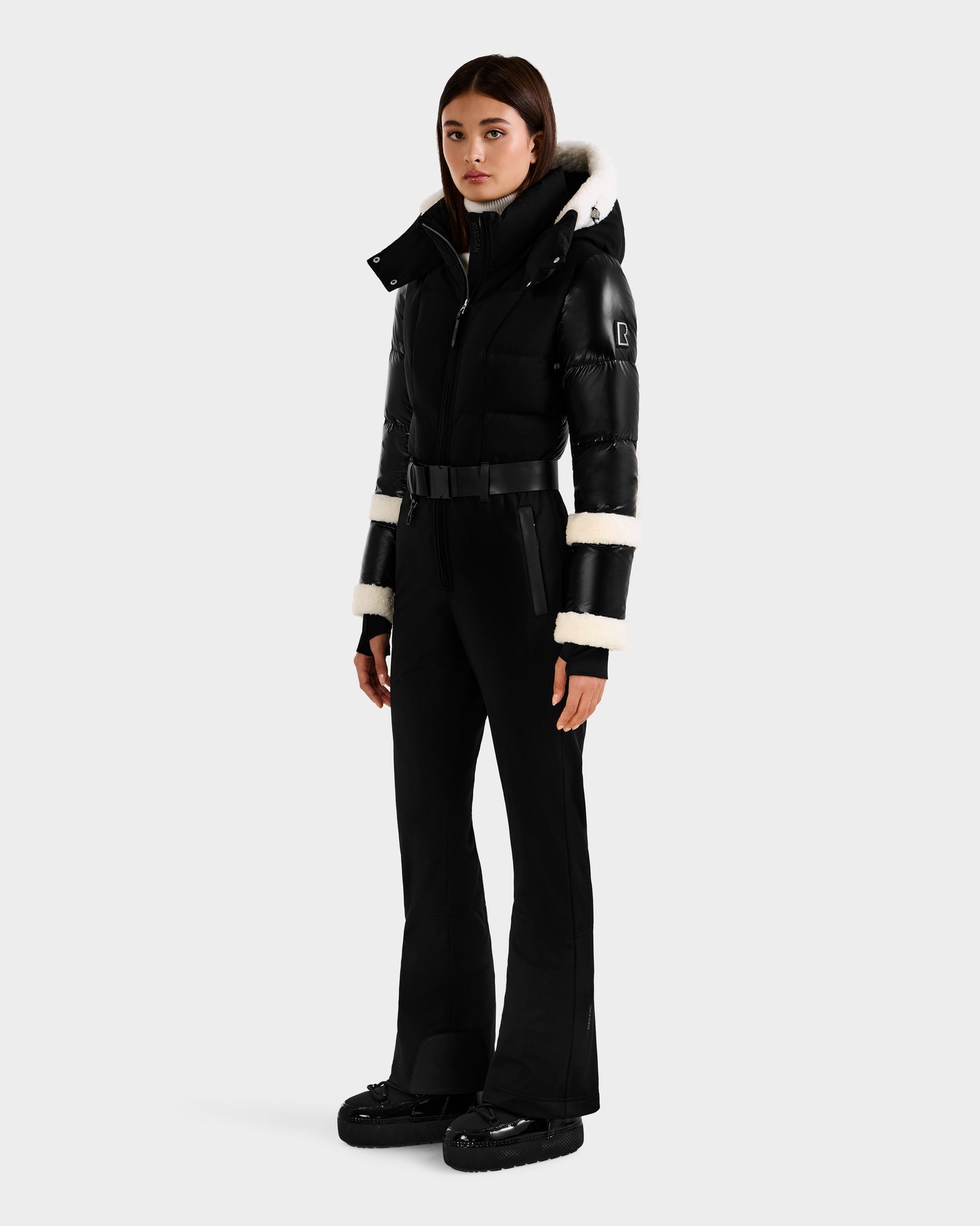 A woman faces forward in the CARMEN - 8125708 Black one-piece ski suit by Rudsak, detailed with white fur on the hood and sleeves, black platform boots, and built-in RECCO rescue tech, set against a plain light gray background.