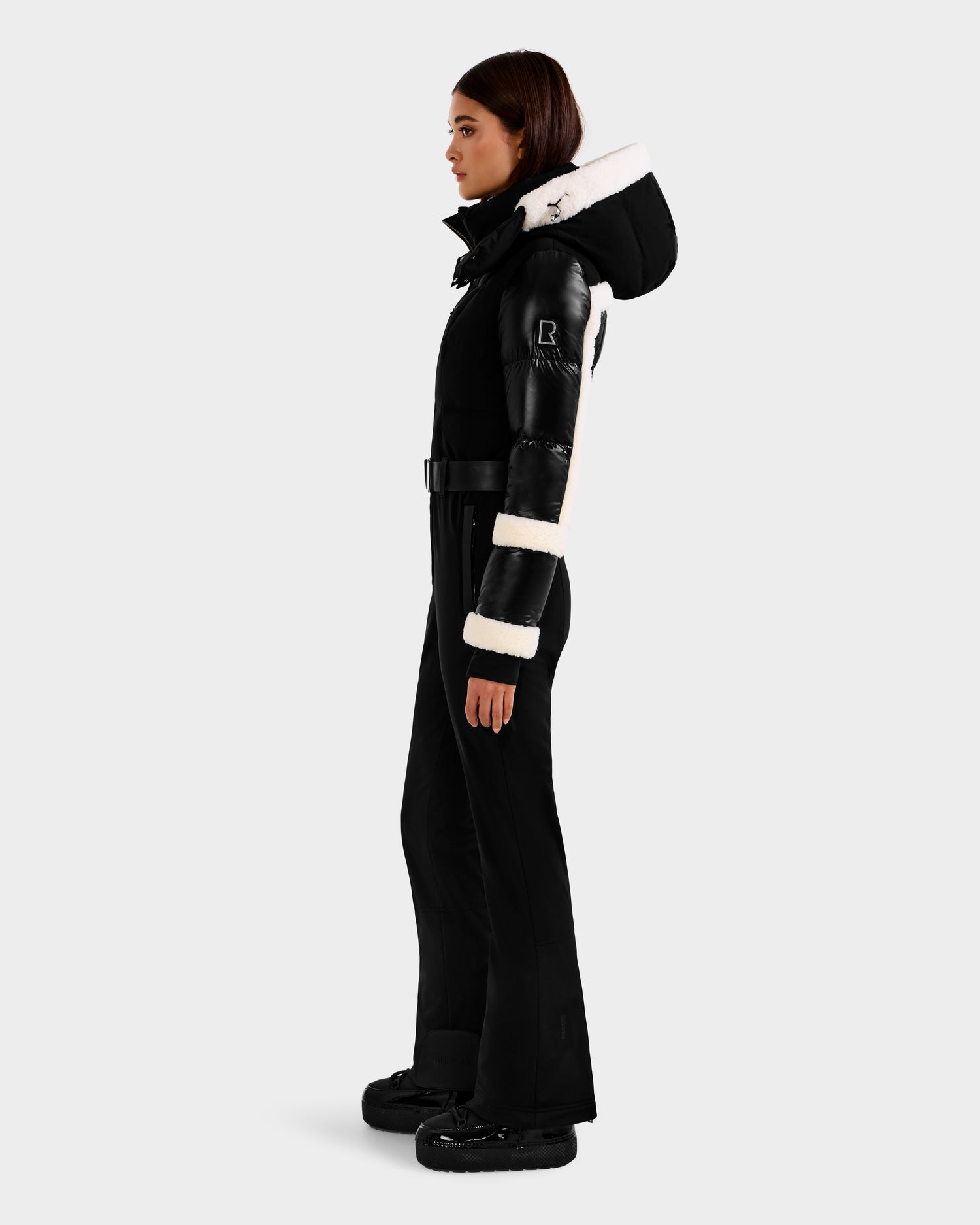 A woman stands in profile wearing the Rudsak CARMEN - 8125708 Black fitted one-piece ski suit, featuring white fur trim on the hood and sleeves, belted at the waist, with black boots against a plain light gray background.