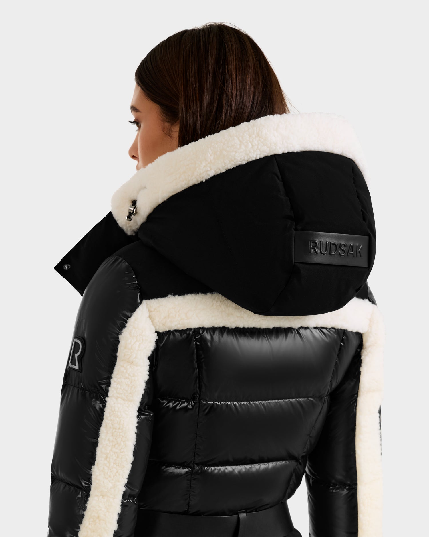 A woman with brown hair and light skin is seen from the back wearing a black Rudsak CARMEN - 8125708 puffer jacket with white fleece trim, a large branded hood, and RECCO rescue technology for safety.