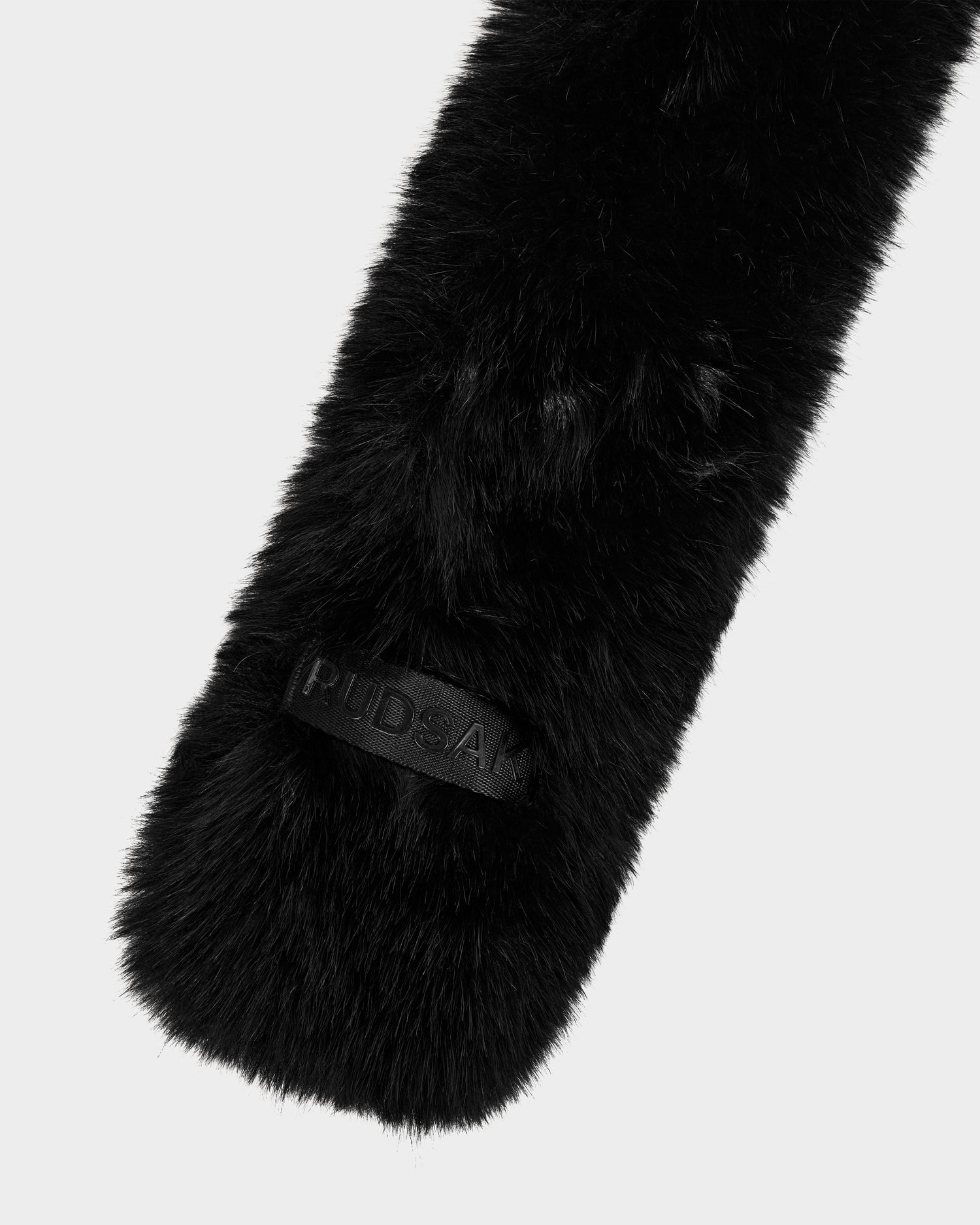 A close-up of the CARMY - L425534 Black by Rudsak, a plush black furry item featuring a rectangular patch embossed with PUDSA, shown against a plain white background.