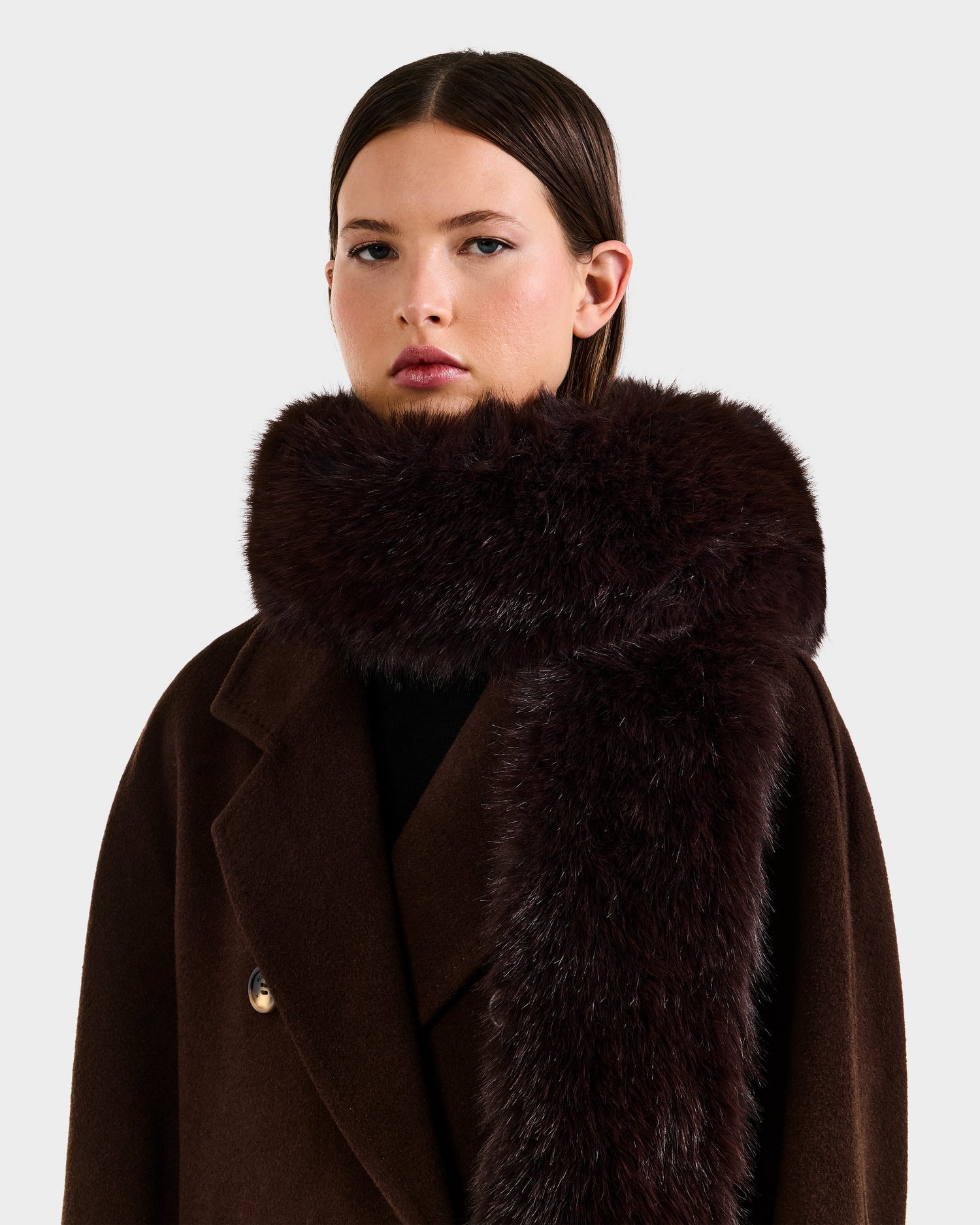 A woman with straight, dark brown hair wears the Rudsak CARMY - L425534 Espresso women’s faux-fur scarf, offering plush warmth against a plain light background.