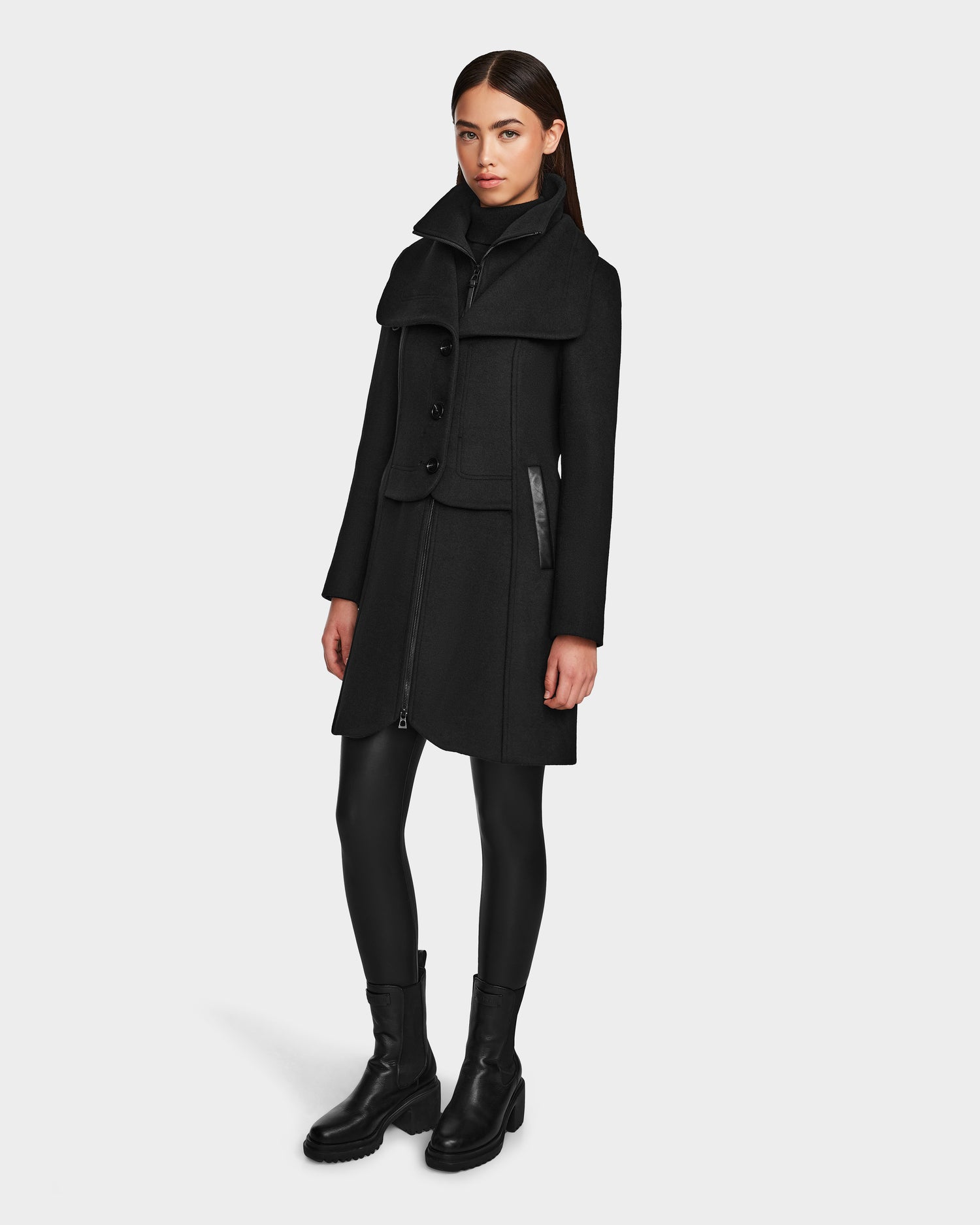 A woman with long brown hair models the Rudsak CAROLINE - L124543 Black knee-length coat, paired with black leggings and ankle boots, standing against a plain white background.