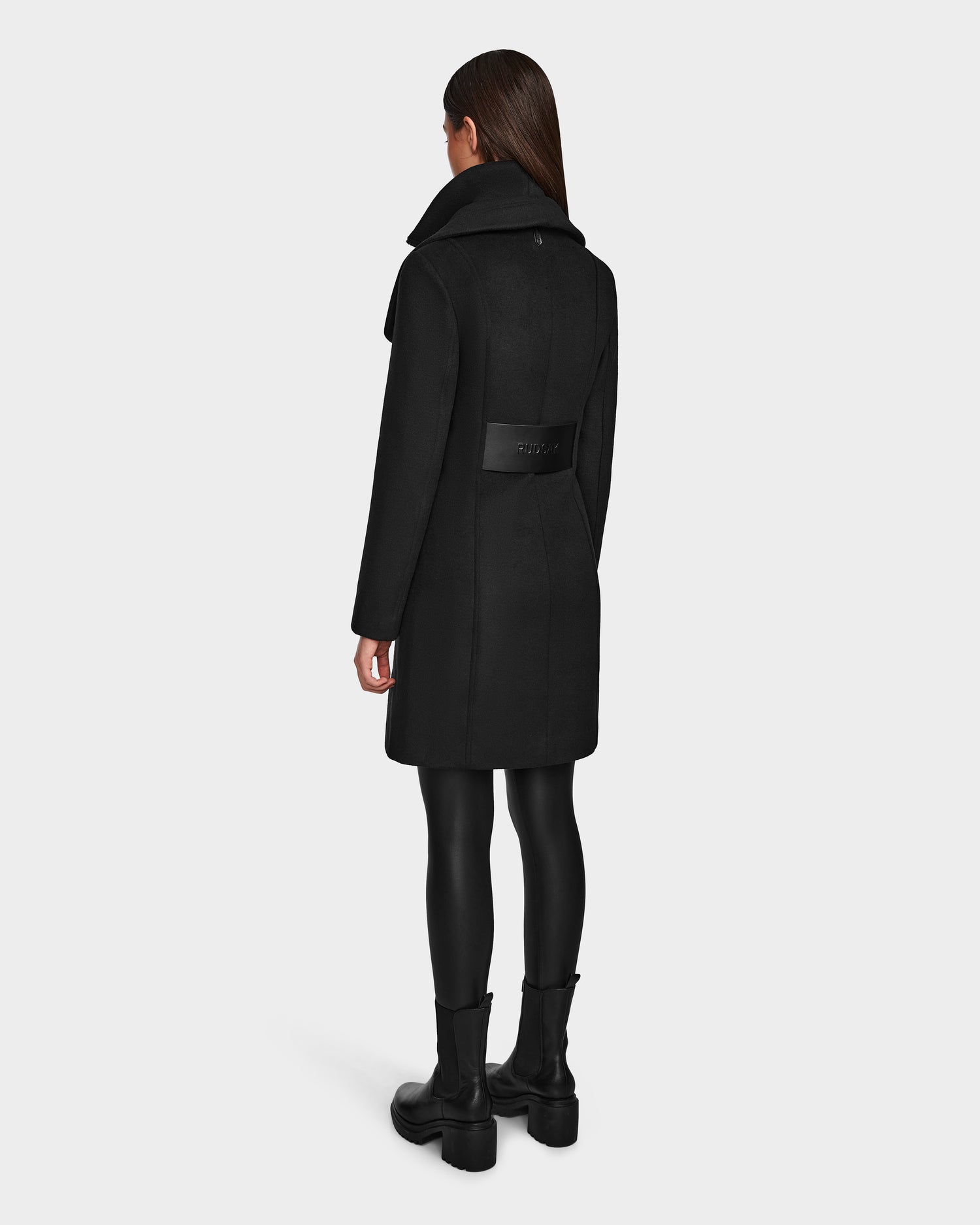A person with long brown hair models the Rudsak CAROLINE - L124543 Black knee-length vegan leather coat, paired with black leggings and boots, facing away to show the back against a plain light gray background.