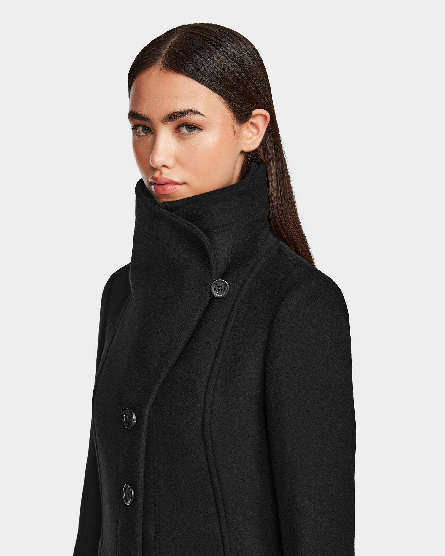 A woman with long, straight brown hair wears the Rudsak CAROLINE - L124543 Black, a high-collared, knee-length premium wool-blend coat with large buttons, standing against a plain light background and looking slightly to the side.