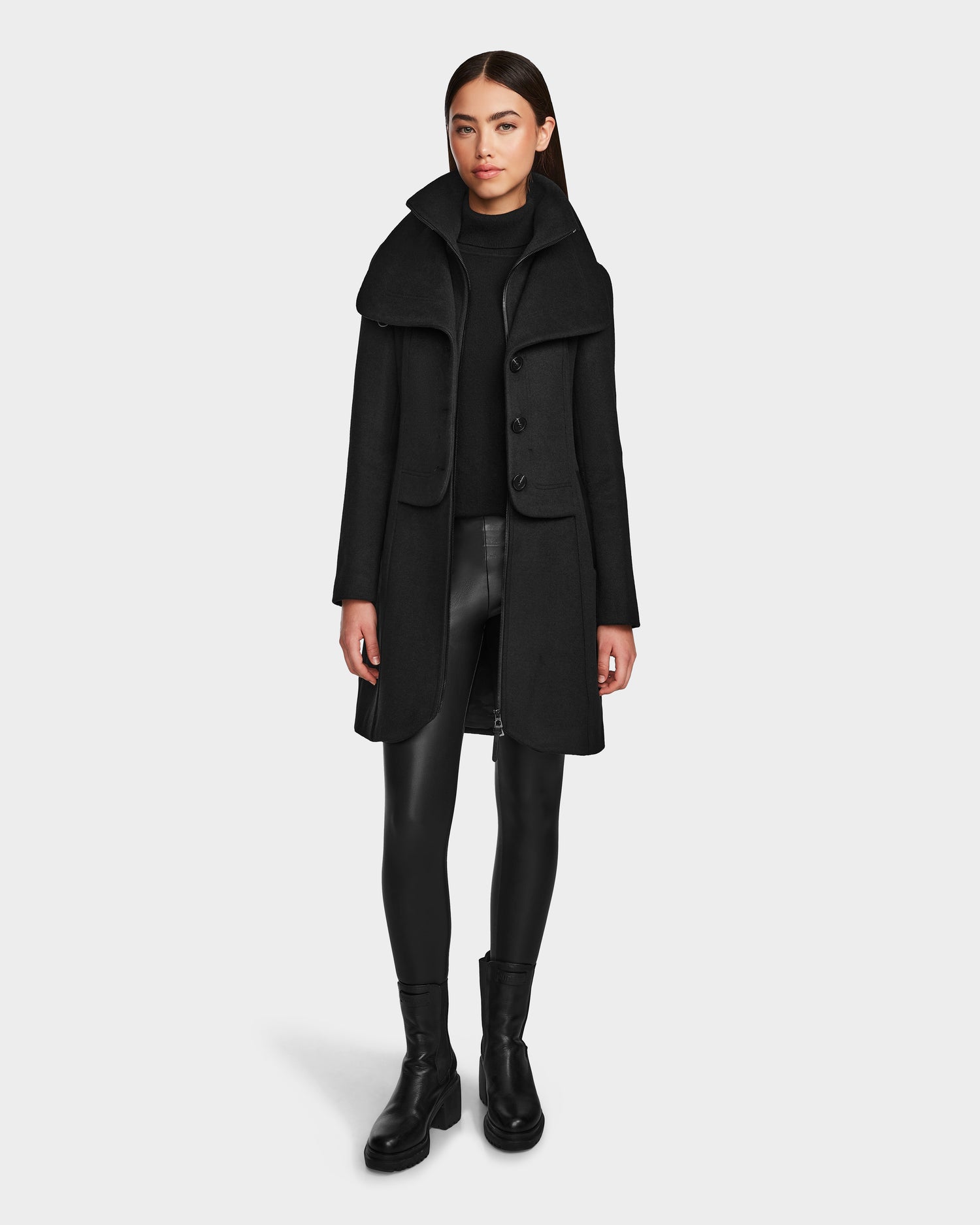 A woman with straight brown hair and a neutral expression models Rudsak’s CAROLINE - L124543 Black: a knee-length, premium wool-blend coat with high collar, styled with a black turtleneck, vegan leather leggings, and ankle boots.