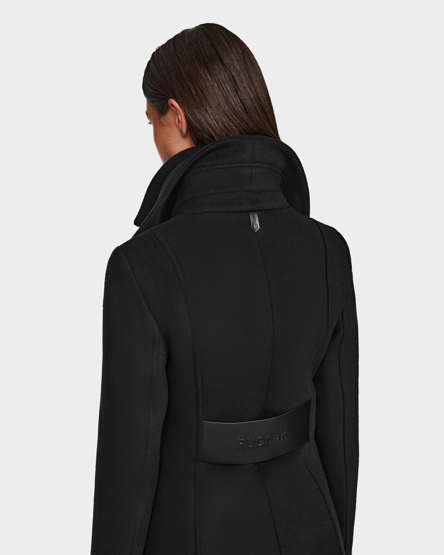 A woman with straight brown hair models the Rudsak CAROLINE - L124543 Black coat, knee-length with a high collar and a back strap embossed with the brand name, photographed from behind.
