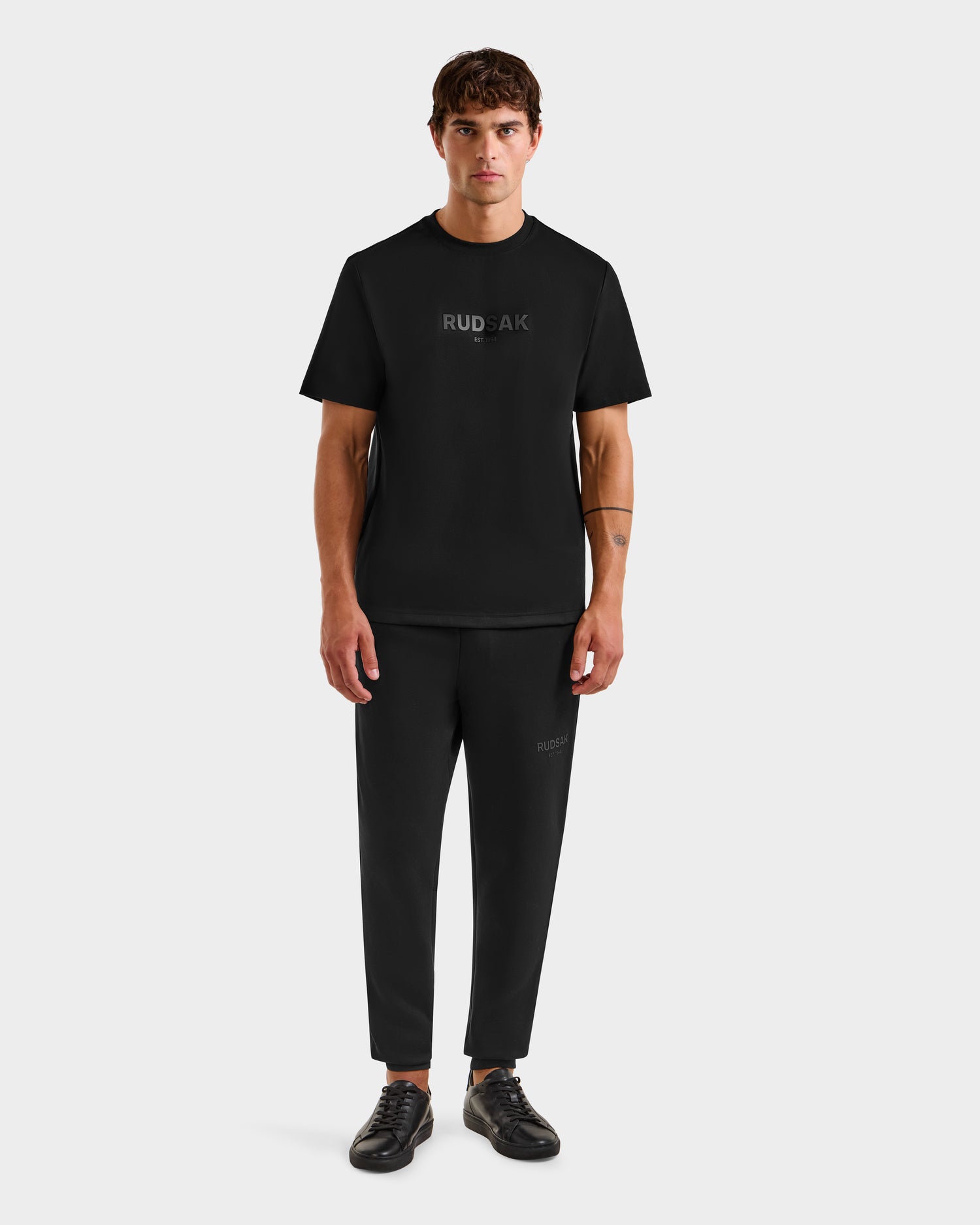 A man stands facing forward in the Rudsak CASEY - 6525037 Black men’s T-shirt, paired with matching logo pants and black sneakers, all set against a plain light gray background.
