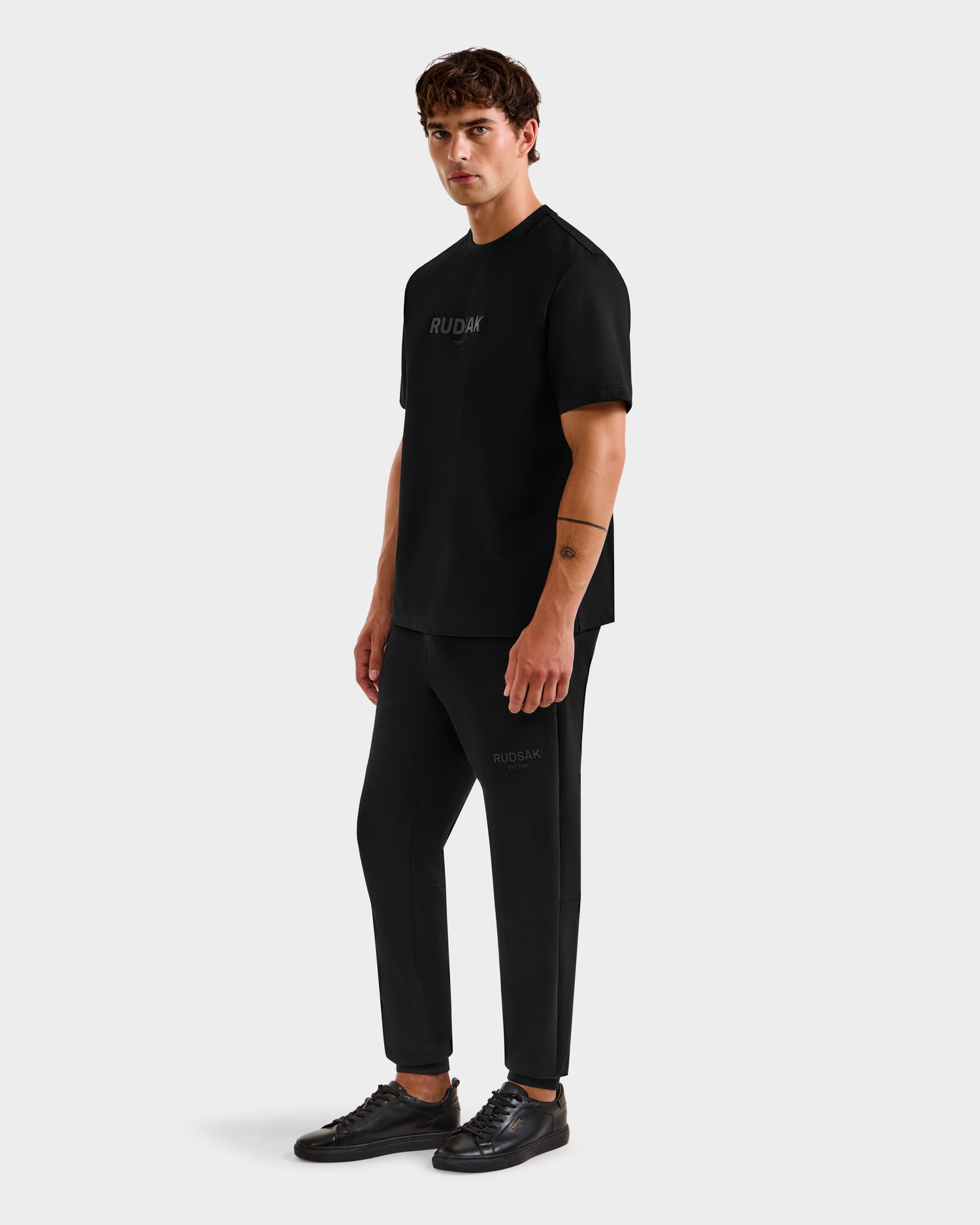 A man with short brown hair and a neutral expression stands against a white background, wearing the Rudsak CASEY - 6525037 Black mens T-shirt, black pants, and black sneakers, showcasing this comfortable essential look.
