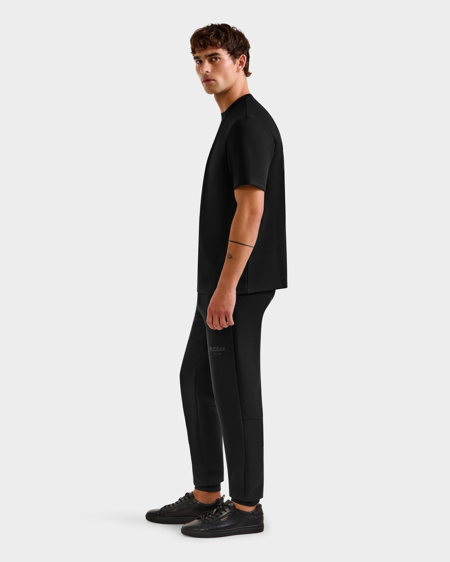 A man with short brown hair and a line tattoo on his left arm stands sideways against a white background, wearing the Rudsak CASEY - 6525037 Black T-shirt, black pants, and black sneakers—showcasing a comfortable essential look.