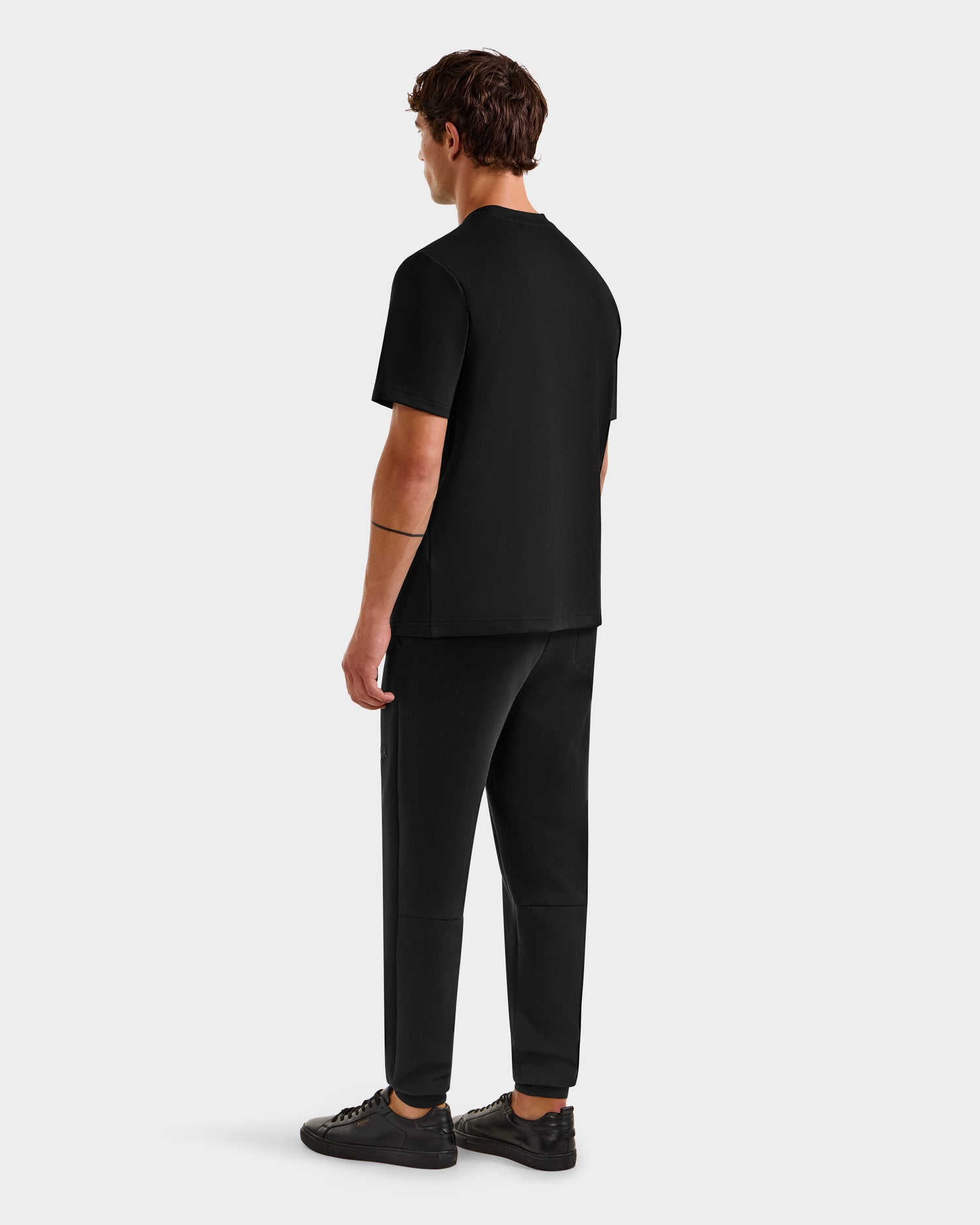 A man faces away from the camera, wearing the Rudsak CASEY - 6525037 Black men’s T-shirt with black pants and sneakers, embodying comfortable essentials against a light gray background.