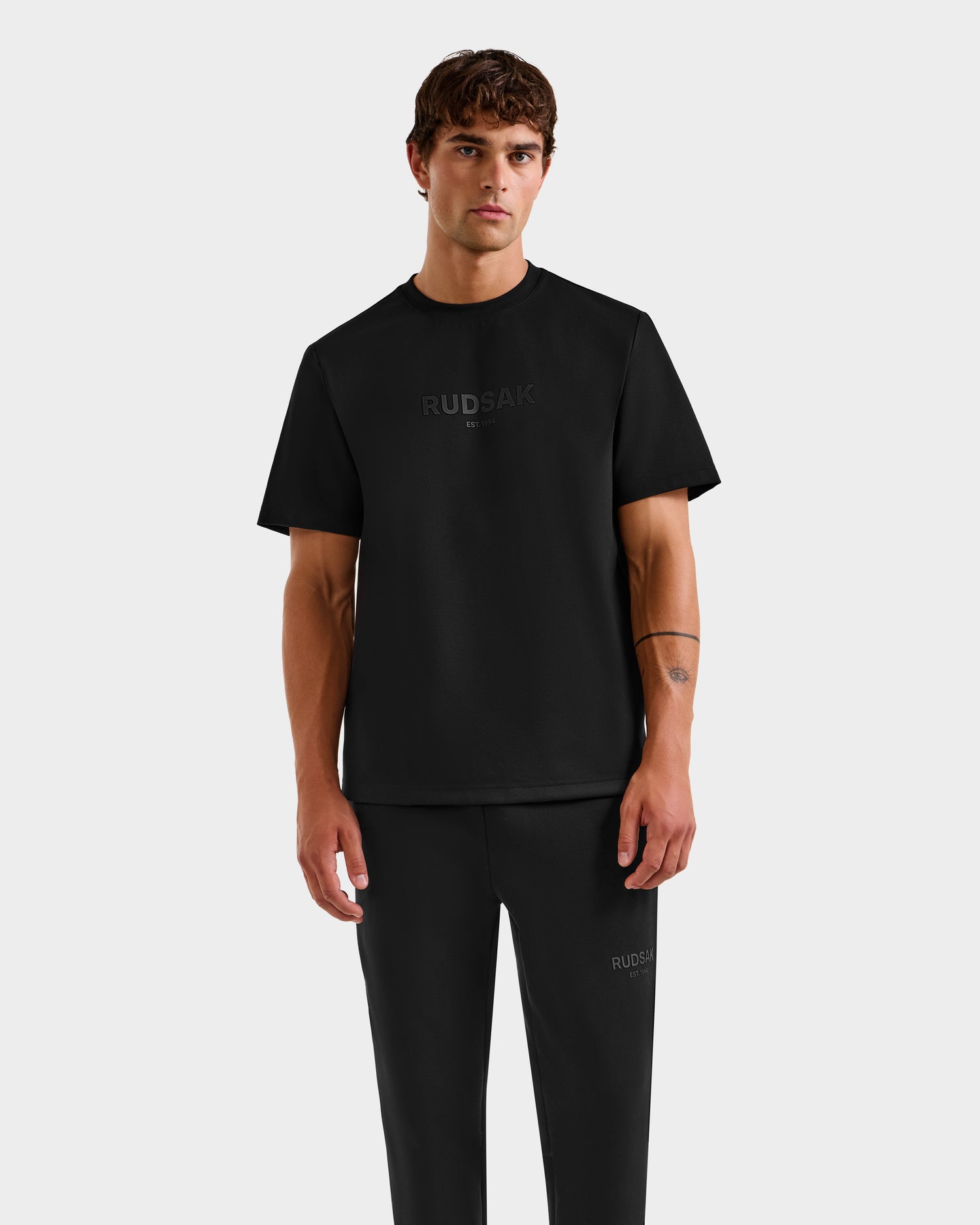 A man with short brown hair wears the Rudsak CASEY - 6525037 Black men's T-shirt and black pants. The shirt features subtle RUDSAK branding on the front. He stands against a plain light gray background, facing forward neutrally.