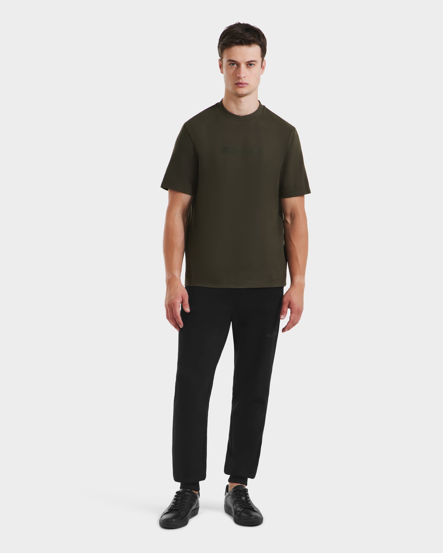 A man faces forward against a plain white background, wearing the Rudsak CASEY - 6525037 dark olive T-shirt with black pants and black sneakers.