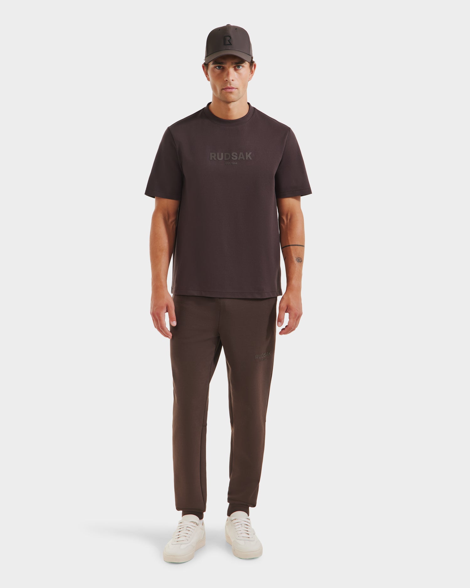 A man stands facing forward in a Rudsak CASEY - 6525037 Espresso T-shirt, matching joggers, a baseball cap, and white sneakers. The dark brown tee features RUDSAK printed on the front against a plain white background.