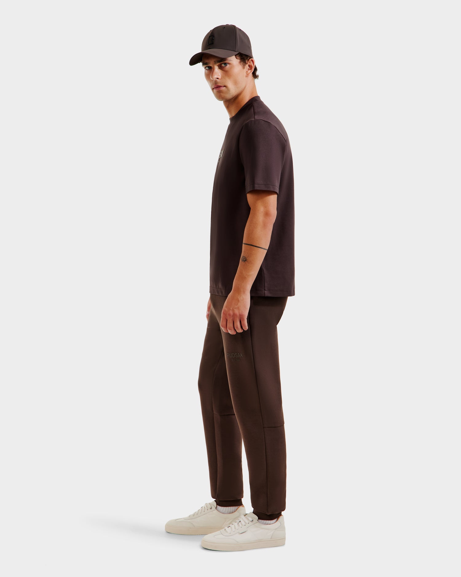A man stands sideways against a plain light background, wearing the Rudsak CASEY - 6525037 Espresso mens T-shirt with matching joggers, a black cap, and white sneakers for a comfortable essential look, gazing toward the camera.
