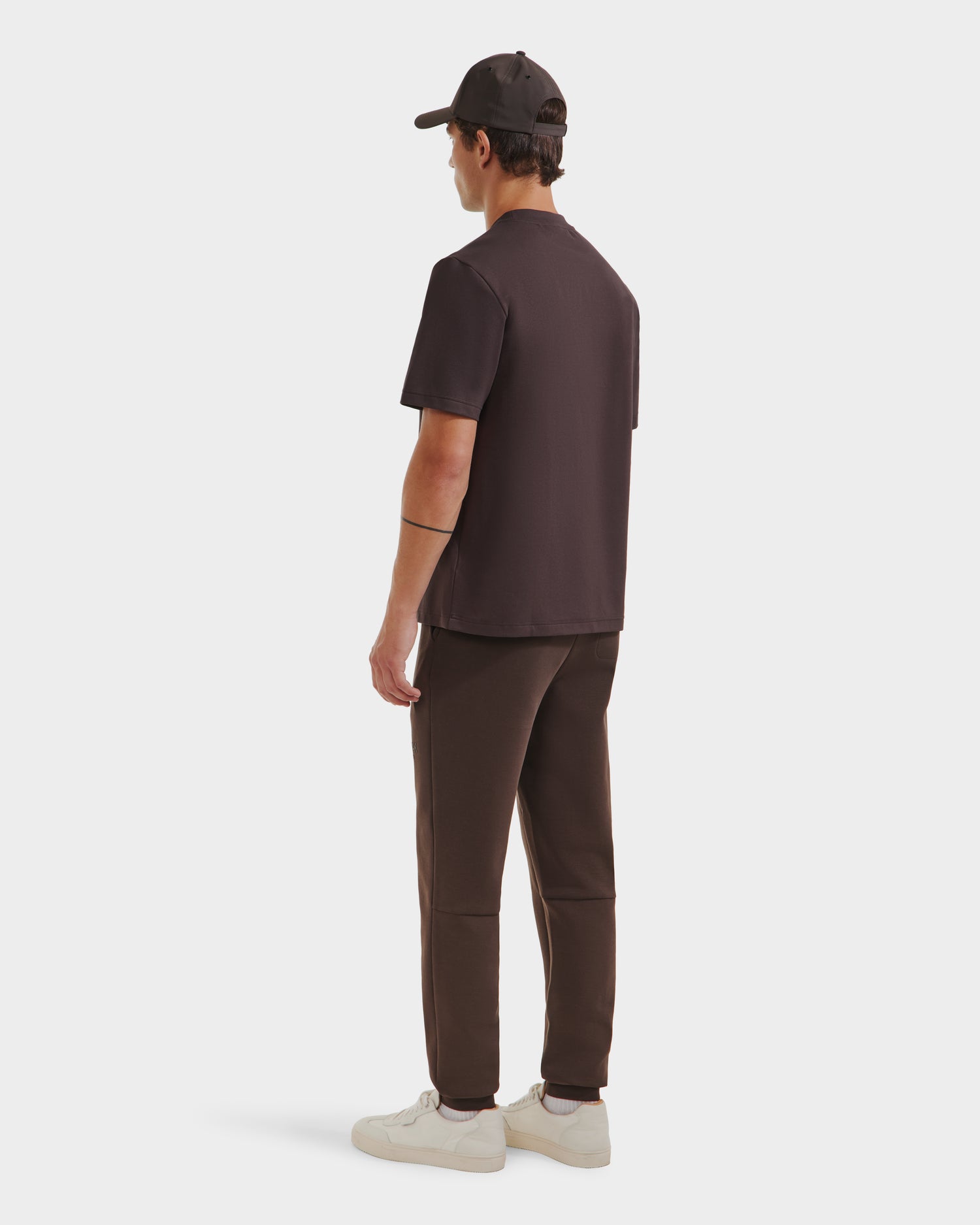 A person faces away, wearing the Rudsak CASEY - 6525037 Espresso mens T-shirt with matching pants, a black baseball cap, and white sneakers against a plain white background.