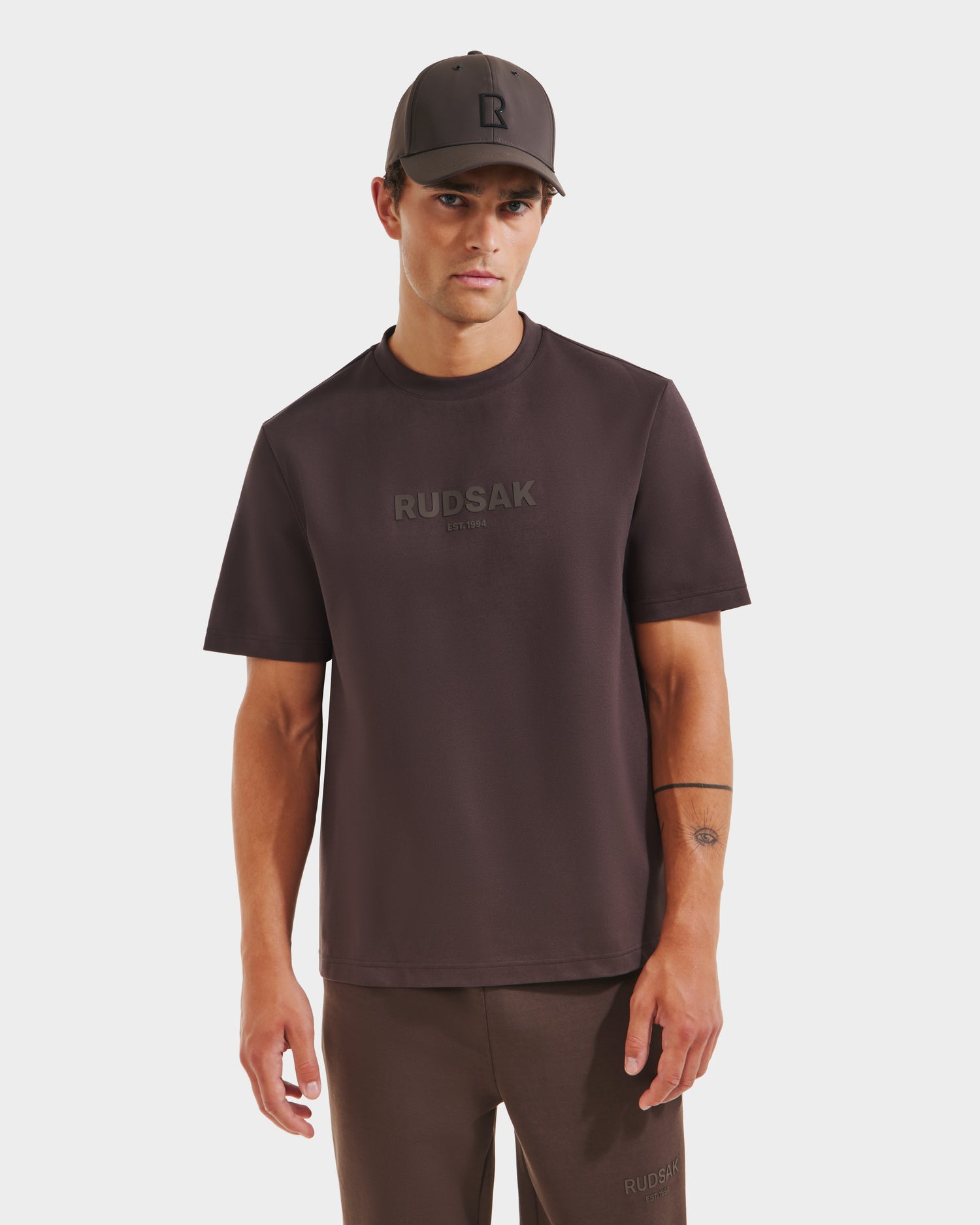 A man wearing a Rudsak CASEY - 6525037 Espresso T-shirt with matching pants and a black cap stands facing forward against a plain white background.