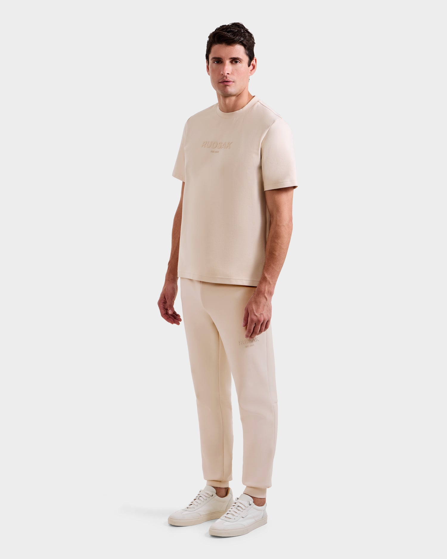 A man stands against a plain background wearing the Rudsak CASEY - 6525037 Vanilla mens T-shirt and matching joggers, paired with white sneakers. He faces slightly to the side with a relaxed expression, highlighting this comfortable essential.