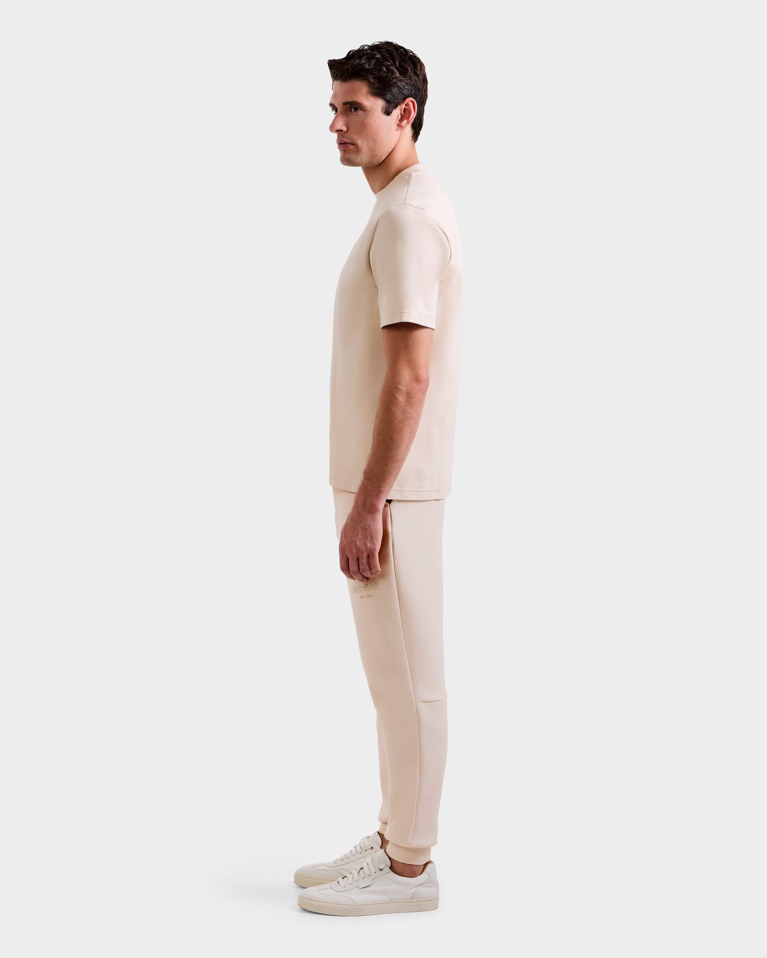 A man stands in profile against a plain background, wearing the Rudsak CASEY - 6525037 Vanilla mens T-shirt and pants set, paired with white sneakers.