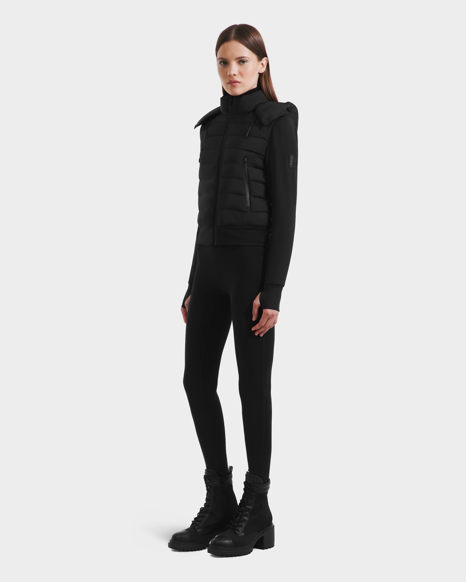 A woman with straight brown hair and a neutral expression stands against a white background, wearing the Rudsak CASSA - 8125057 Black lightweight cropped jacket over a fitted black outfit and lace-up boots—ideal for transitional weather.