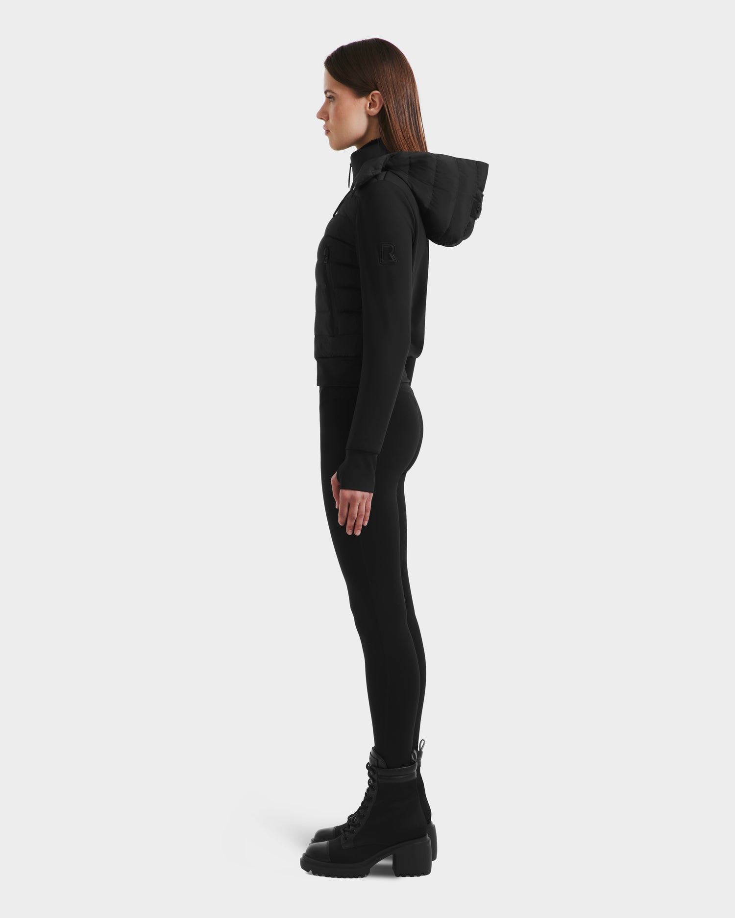 A woman stands in profile facing left, dressed in Rudsak’s CASSA - 8125057 Black: an all-black, lightweight cropped jacket, paired with fitted pants and chunky lace-up boots, set against a plain white background.