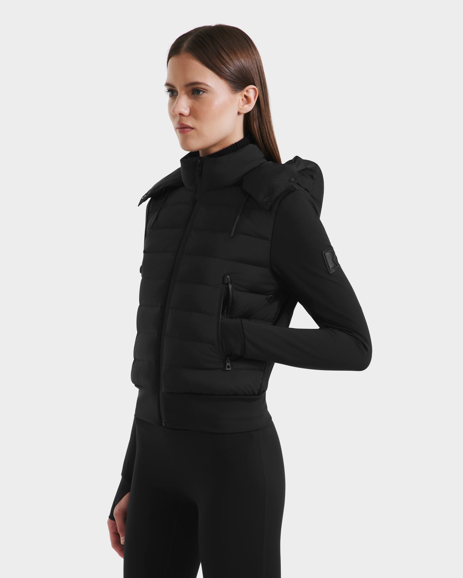 A woman with straight brown hair models the Rudsak CASSA - 8125057 Black fitted women’s jacket with a high collar and zippered pockets, layered over black attire, standing against a plain white background.