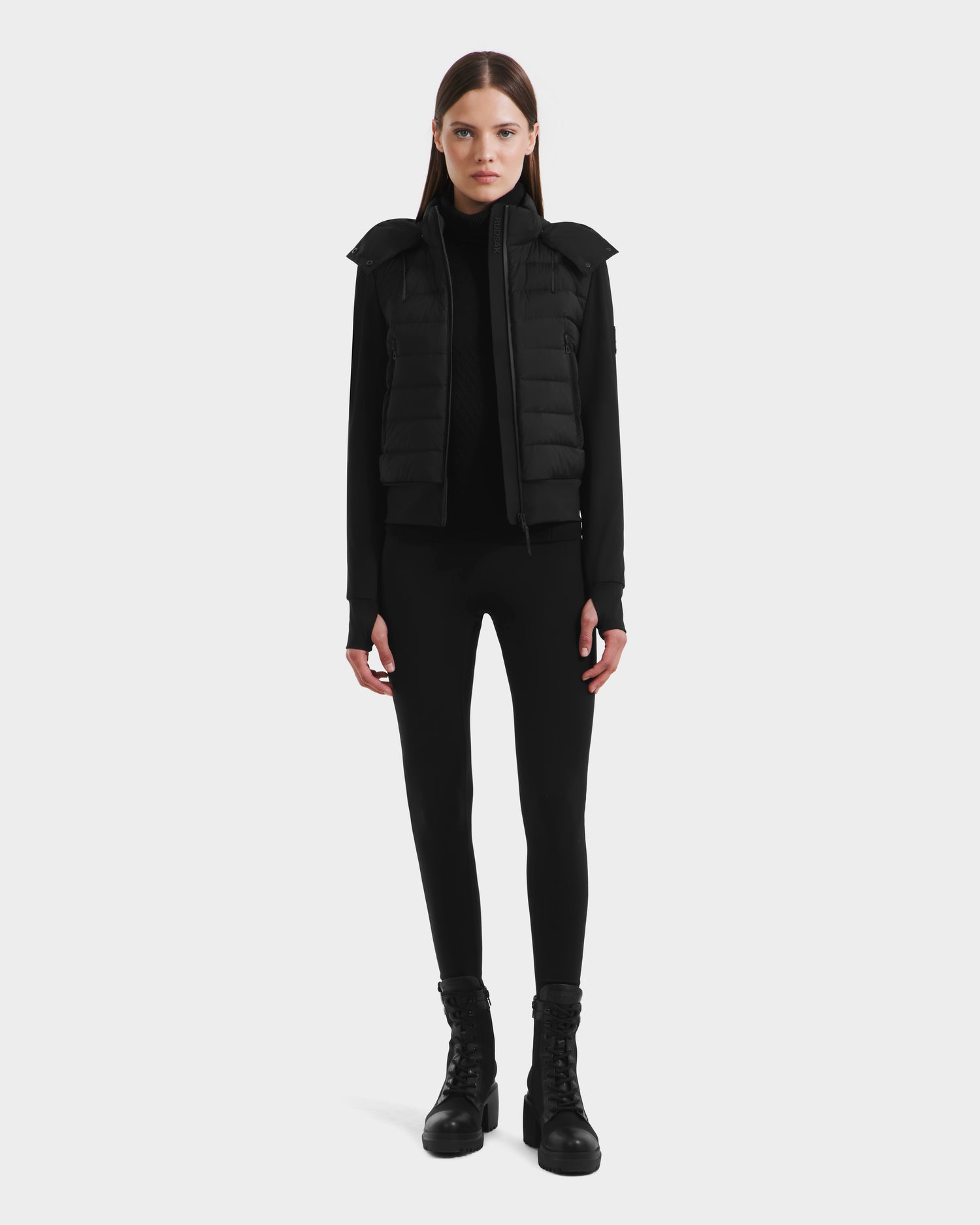 A woman stands facing forward in the Rudsak CASSA - 8125057 Black lightweight cropped jacket, layered over a black top, leggings, and lace-up boots—an ideal transitional weather look—set against a plain white background.
