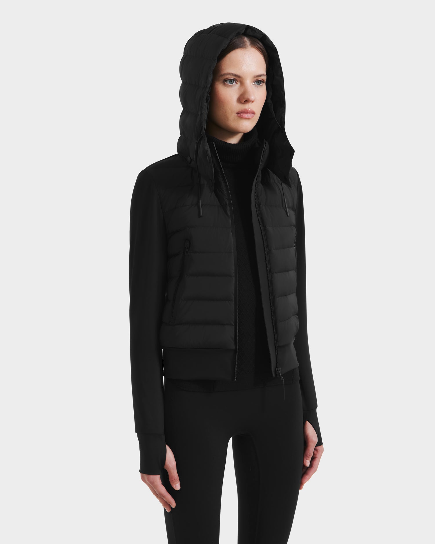 A woman faces slightly to the side, wearing the Rudsak CASSA - 8125057 Black lightweight cropped jacket over black leggings—a chic pick for transitional weather—against a plain white background.