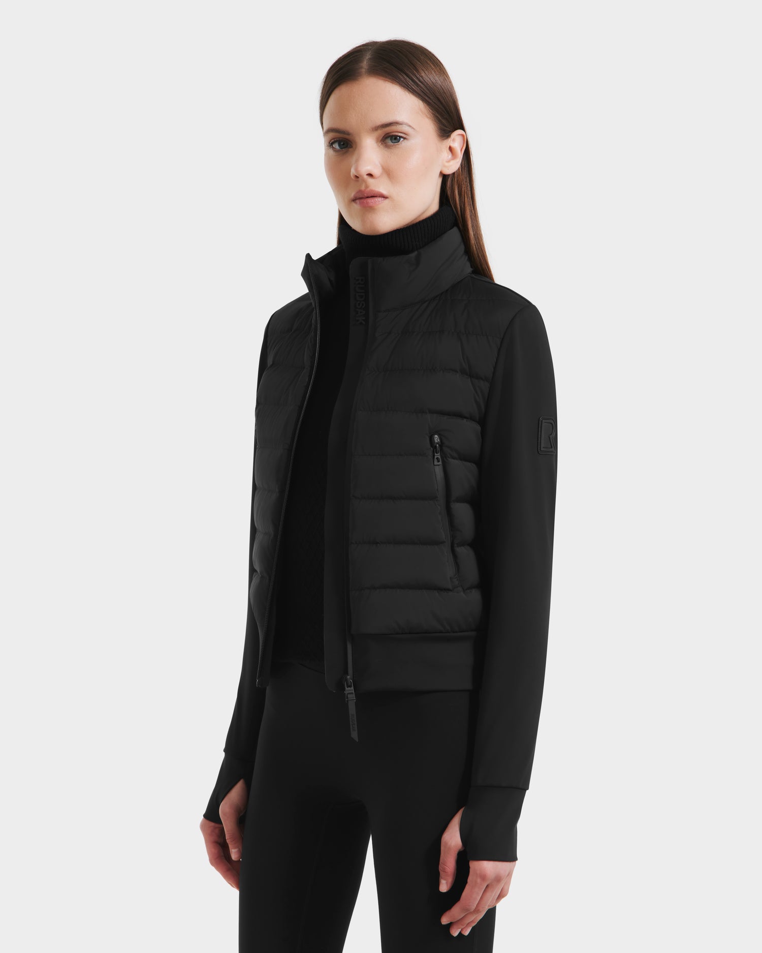 A woman with straight brown hair models Rudsak’s CASSA - 8125057 Black, a lightweight cropped puffer jacket with a high collar, styled over a black turtleneck and fitted pants—ideal for transitional weather against a light backdrop.