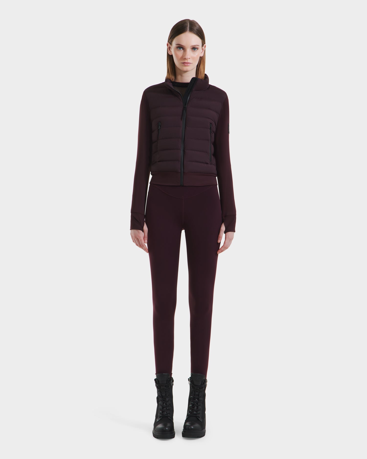 A woman faces forward against a white background, wearing the Rudsak CASSA - 8125057 Bordeaux transitional jacket with padded panels, paired with matching high-waisted leggings and black lace-up boots.