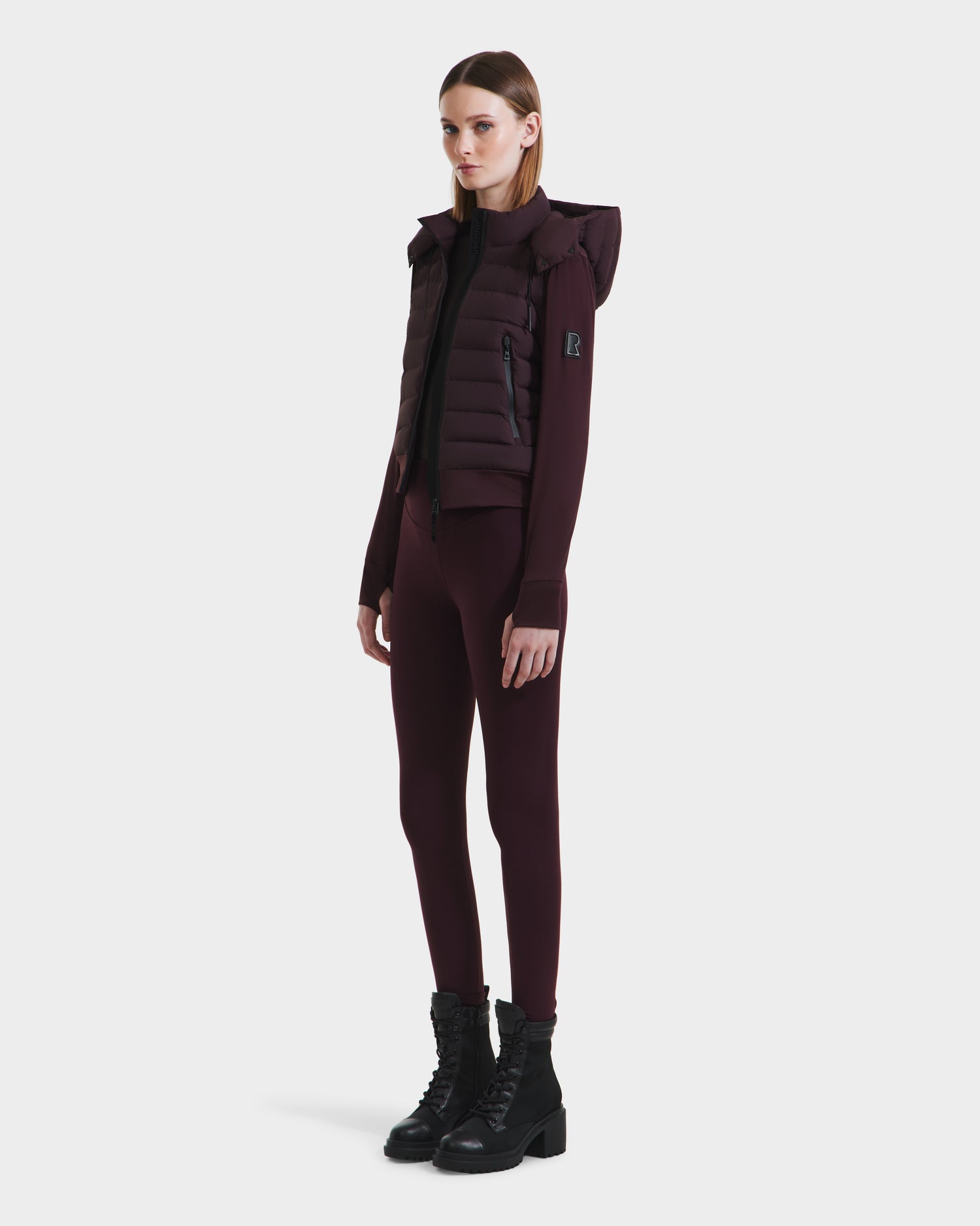 A woman poses confidently in the CASSA - 8125057 Bordeaux transitional jacket by Rudsak, layered over a long-sleeve top and leggings, with black lace-up boots, against a plain white background.