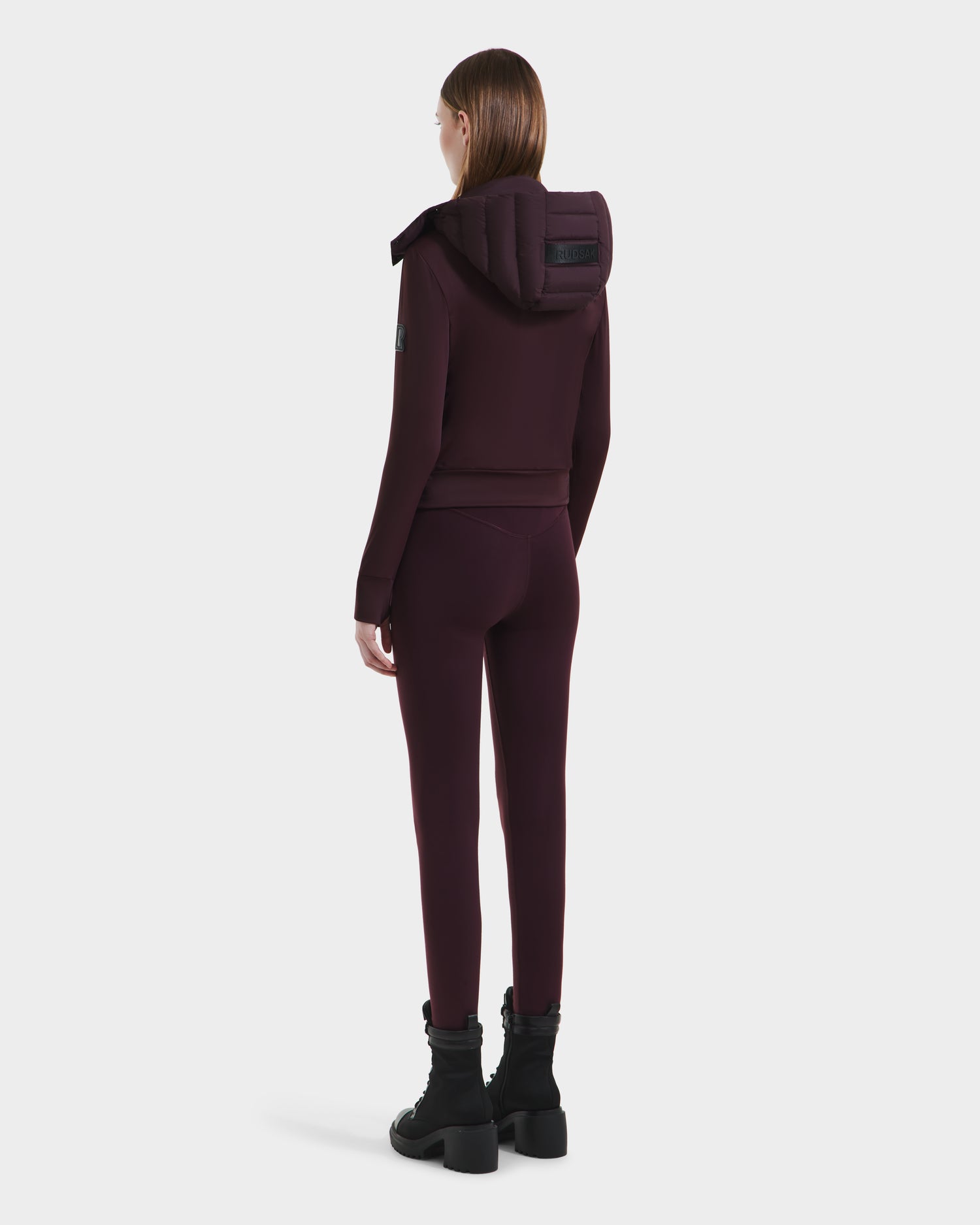 A woman faces away, wearing the Rudsak CASSA - 8125057 Bordeaux set—matching transitional jacket and fitted pants—with black ankle boots, shown against a plain white background.