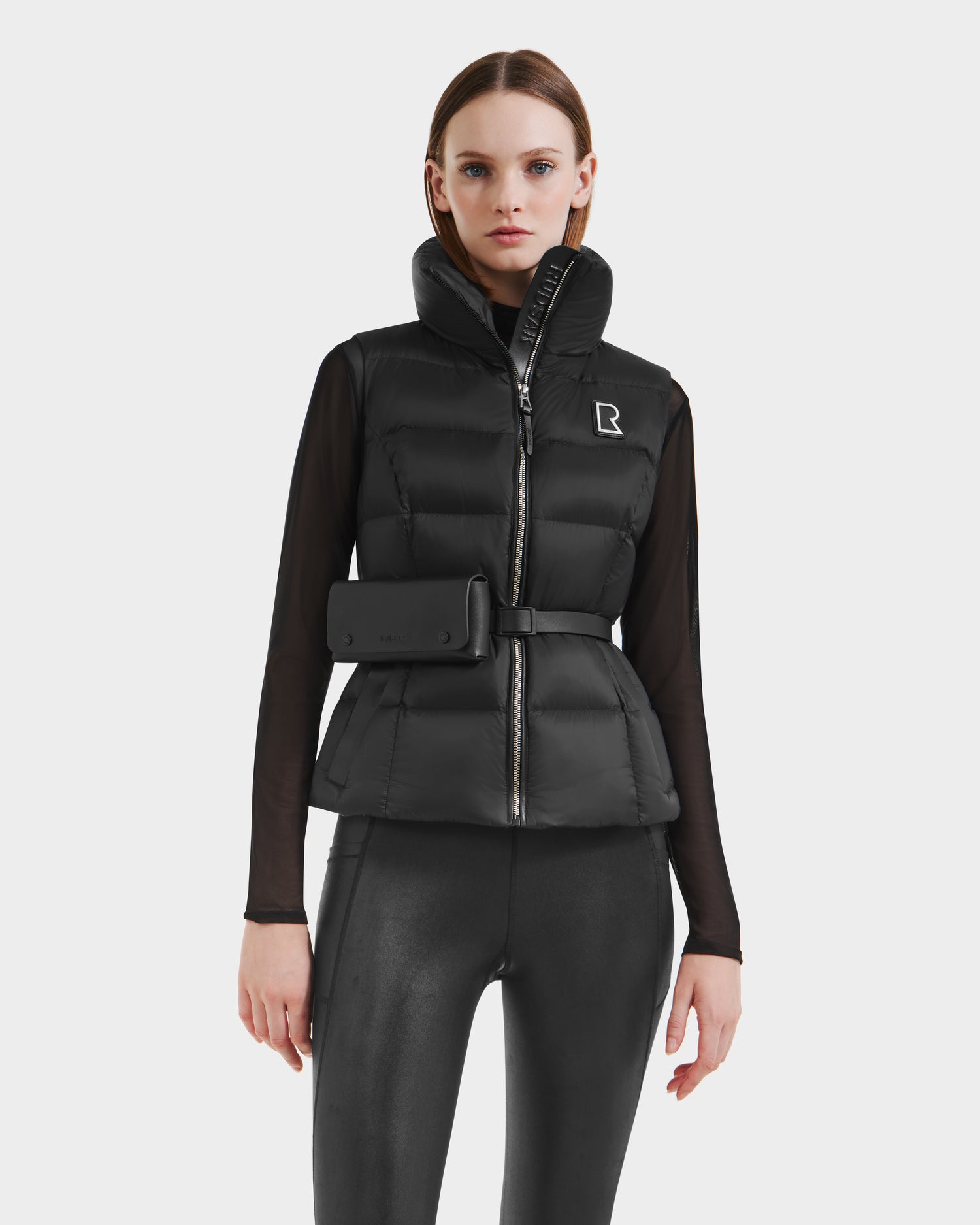 A woman wears the Rudsak CASSIDY N - 8125677 Black recycled nylon fitted down vest, styled with a black belt bag, sheer-sleeved top, and shiny fitted pants, posing against a plain white background.
