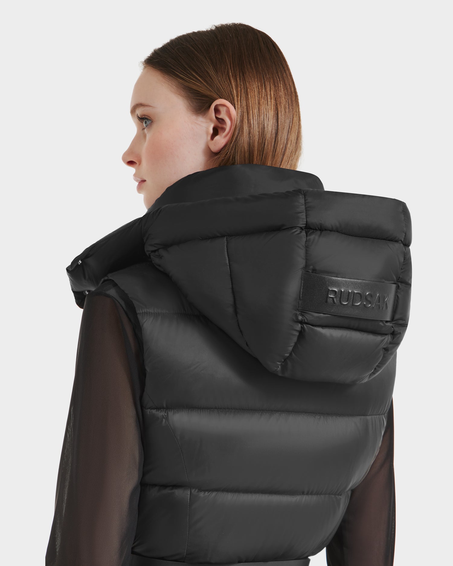 A woman with straight light brown hair models the Rudsak CASSIDY N - 8125677 Black women’s fitted down vest with a large hood and visible branding, shown in profile against a plain background to highlight its lightweight, travel-friendly warmth.