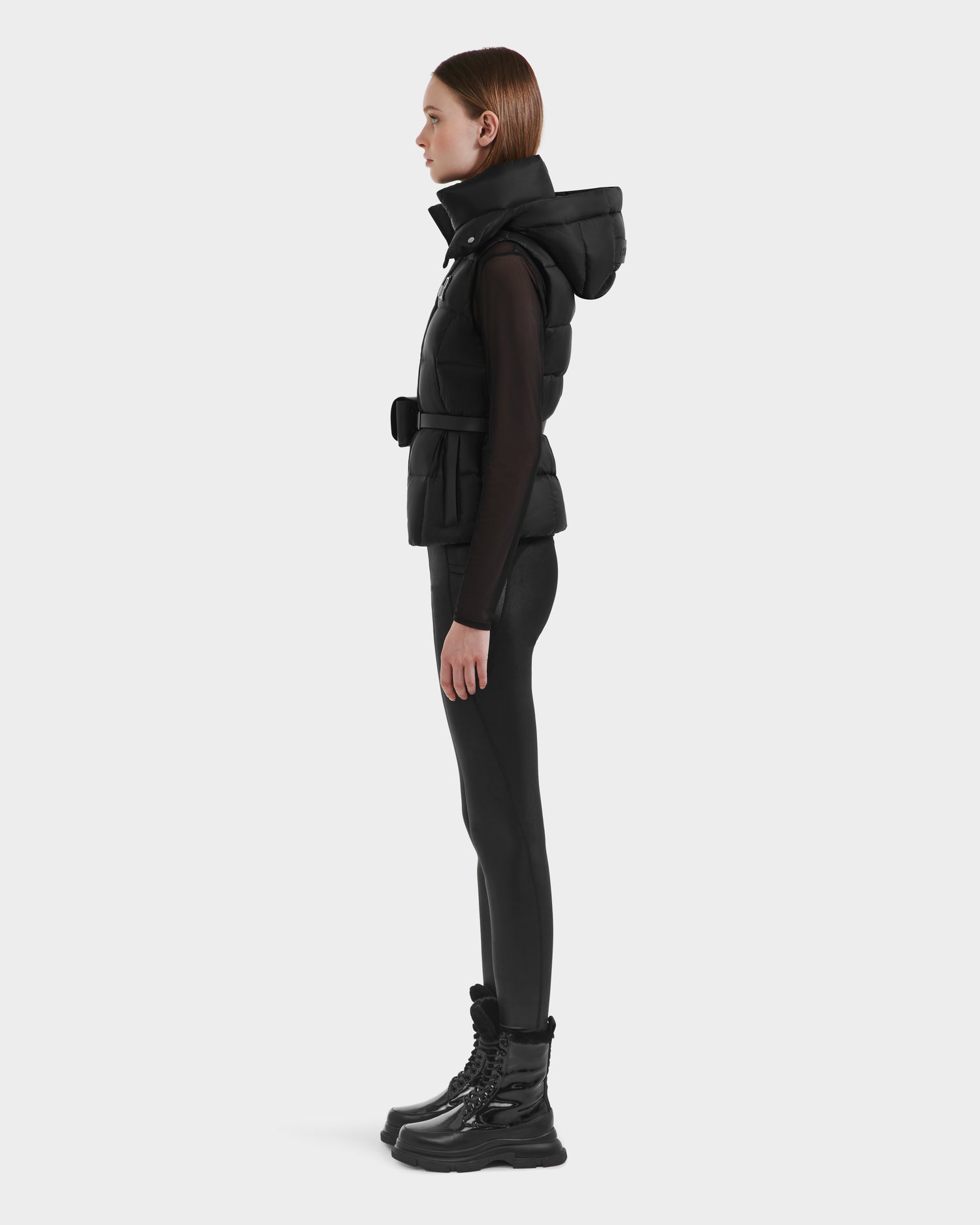 A woman in profile wears the Rudsak CASSIDY N - 8125677 Black women’s fitted down vest, paired with a black long-sleeve top, leggings, and lace-up boots, standing against a plain light background.