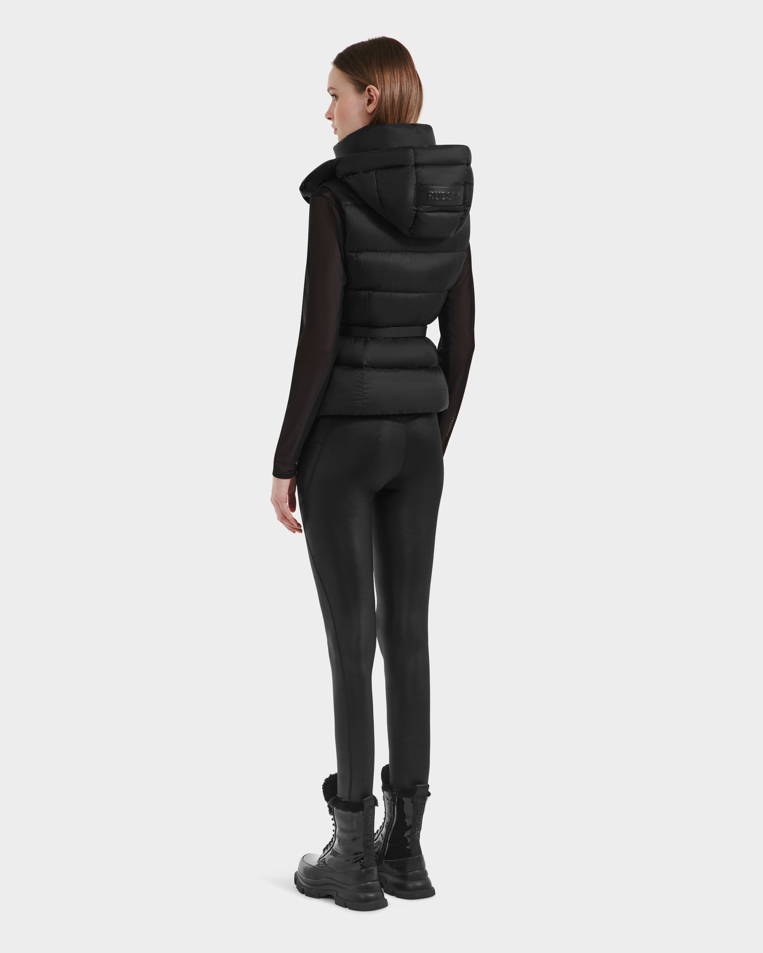 A woman, seen from behind, wears the Rudsak CASSIDY N - 8125677 Black fitted down vest with hood over a black long-sleeve top, leggings, and lace-up boots—an ideal travel-friendly layer against a plain white background.
