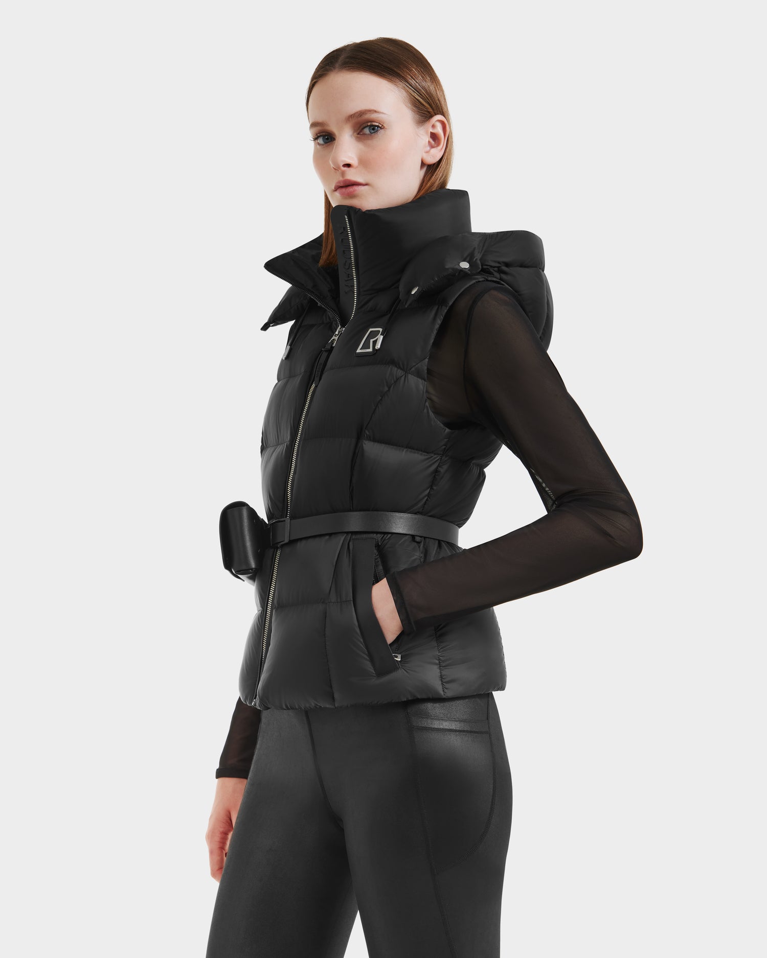 A woman wears the Rudsak CASSIDY N - 8125677 Black women’s fitted down vest with a belt over a sheer black top and fitted pants, confidently showcasing this travel-friendly layer against a plain light background.