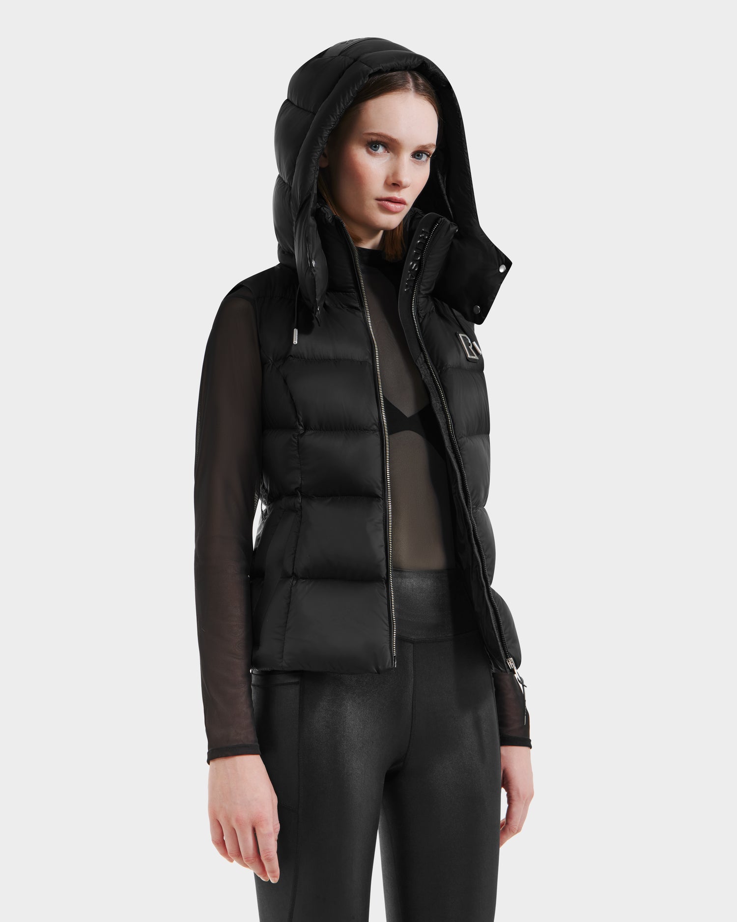 A woman models the Rudsak CASSIDY N - 8125677 Black fitted down vest with hood over a sheer black top and leggings against a light gray background, highlighting this travel-friendly layer’s lightweight warmth.