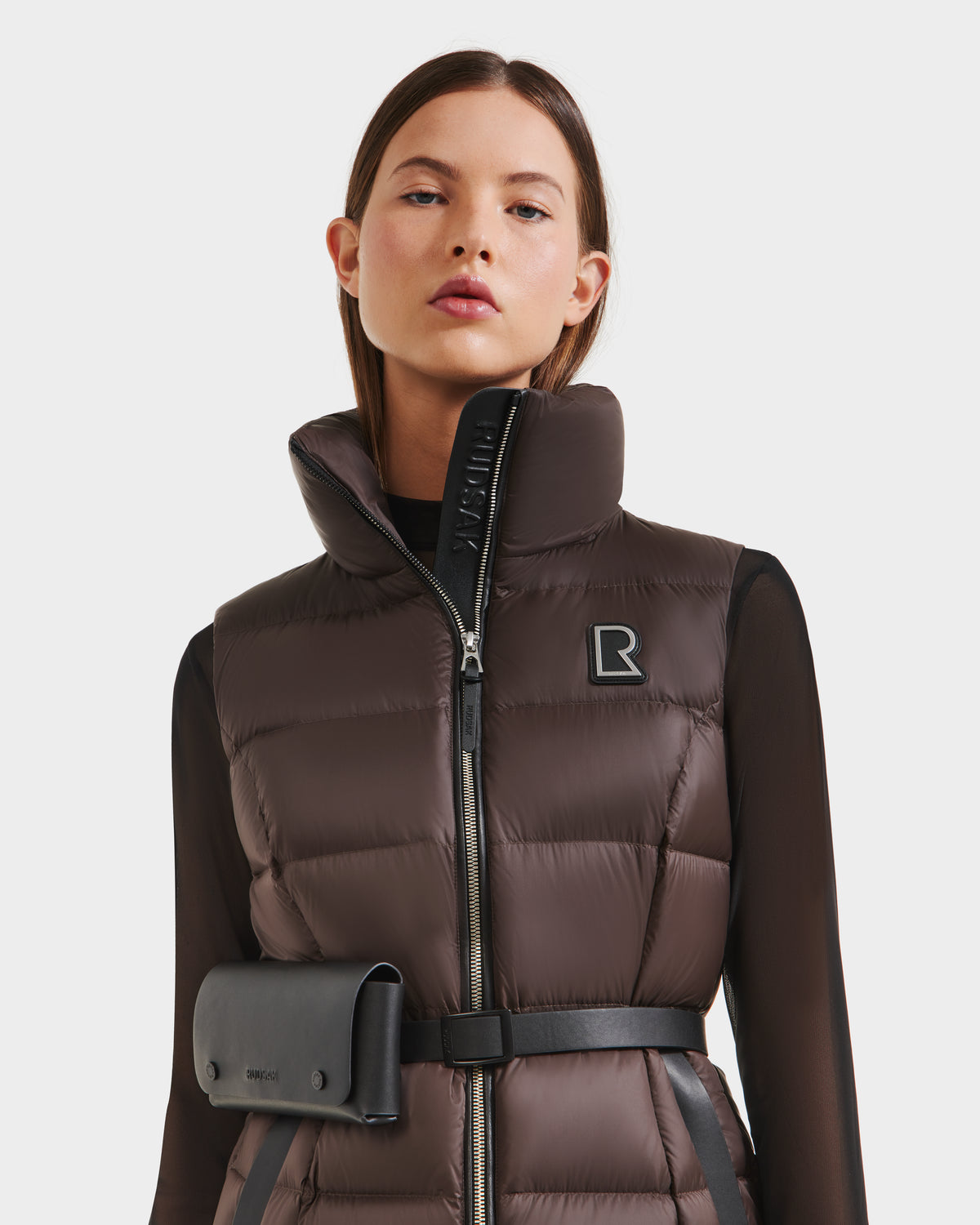 A woman wears the Rudsak CASSIDY N - 8125677 Espresso vest, a brown quilted puffer with lightweight warmth, high collar, black logo patch, and black belt with pouch—perfect for travel-friendly style against a white background.