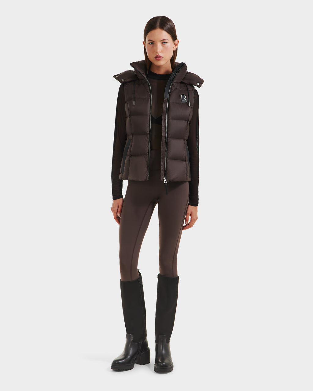 A woman faces forward wearing the Rudsak CASSIDY N - 8125677 Espresso womens fitted down vest for lightweight warmth over a black long-sleeve top, brown leggings with black side panels, and knee-high boots, styled for travel-ready comfort.