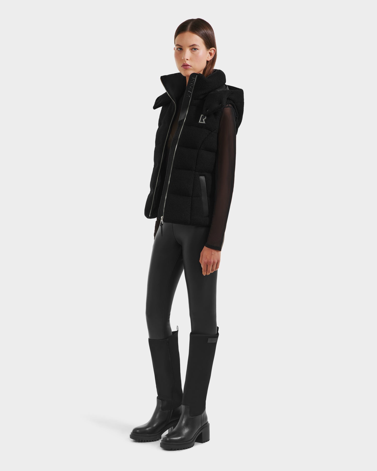A woman with straight brown hair and a neutral expression models the Rudsak CASSIDY - 8125622 Black fitted down vest over a sheer long-sleeve top, paired with black fitted pants and knee-high boots against a plain white background.