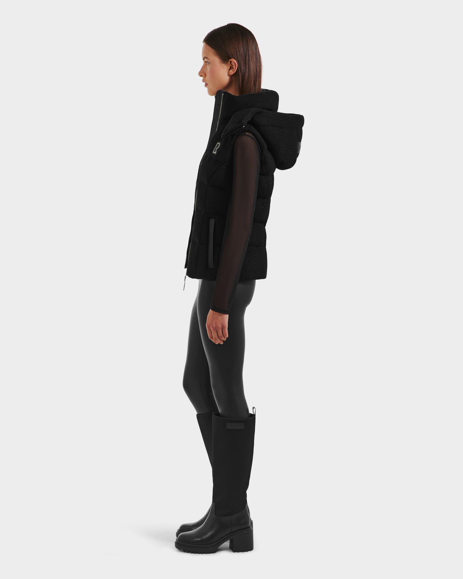 A woman stands in profile wearing the Rudsak CASSIDY - 8125622 Black fitted down vest with a removable hood, paired with a sheer black long-sleeve top, shiny leggings, and knee-high boots—showcasing lightweight warmth on a white background.