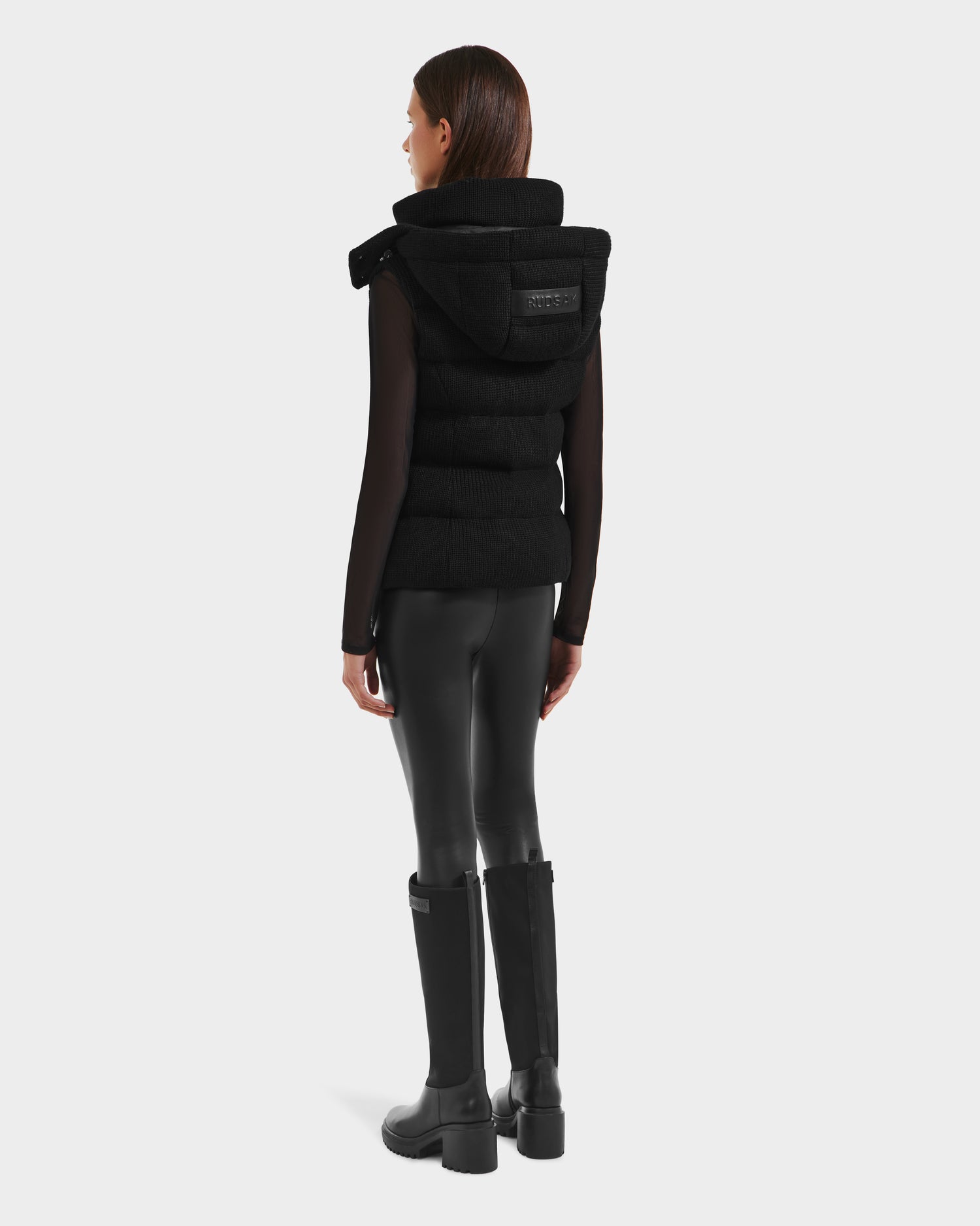 A woman faces away, wearing the Rudsak CASSIDY - 8125622 Black women’s fitted down vest with removable hood, paired with a black top, shiny black leggings, and knee-high boots—merging lightweight warmth and sleek style.