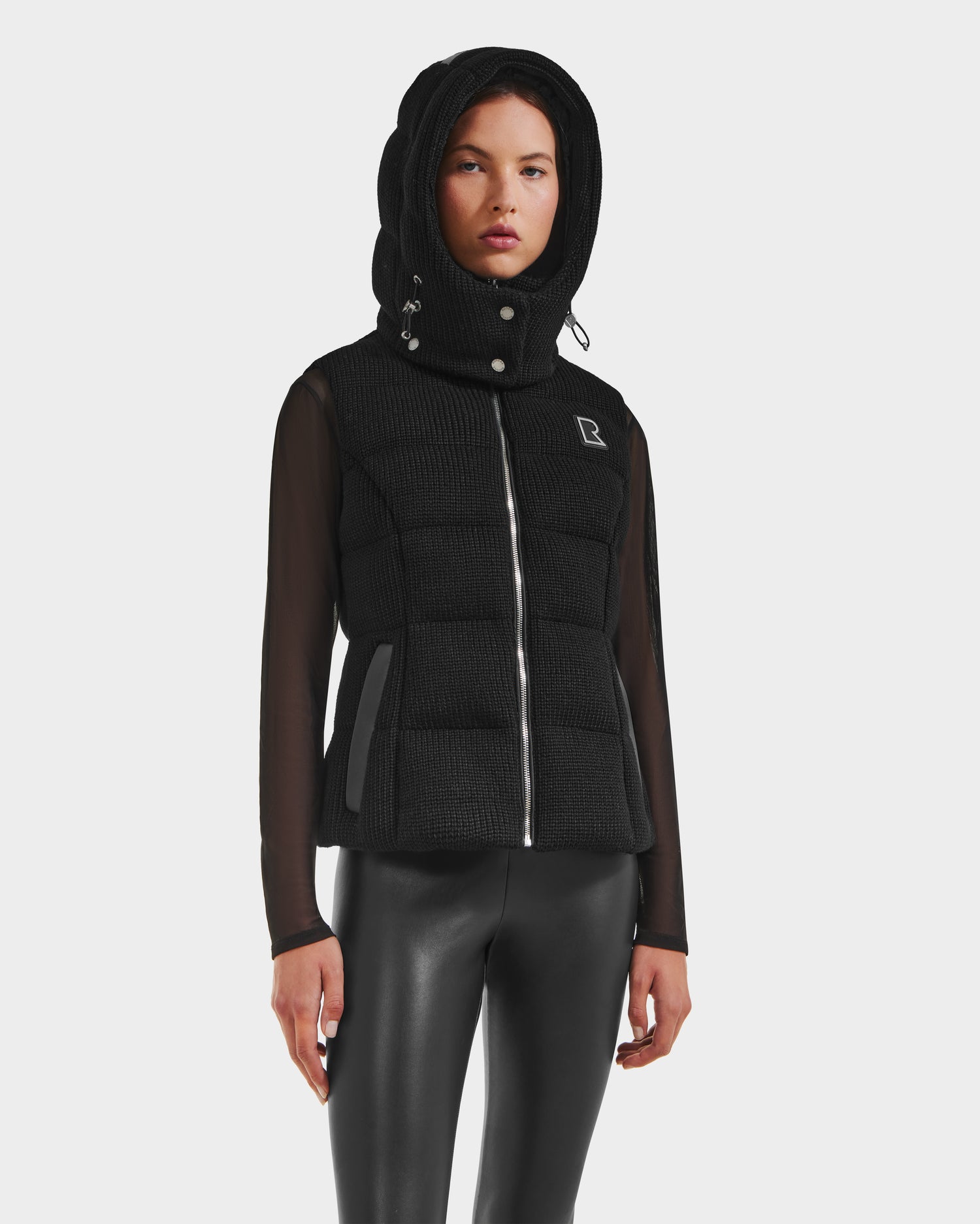 A person wears the Rudsak CASSIDY - 8125622 Black womens fitted down vest with a removable hood, styled with sheer long sleeves and black leather-look pants, against a plain light background.