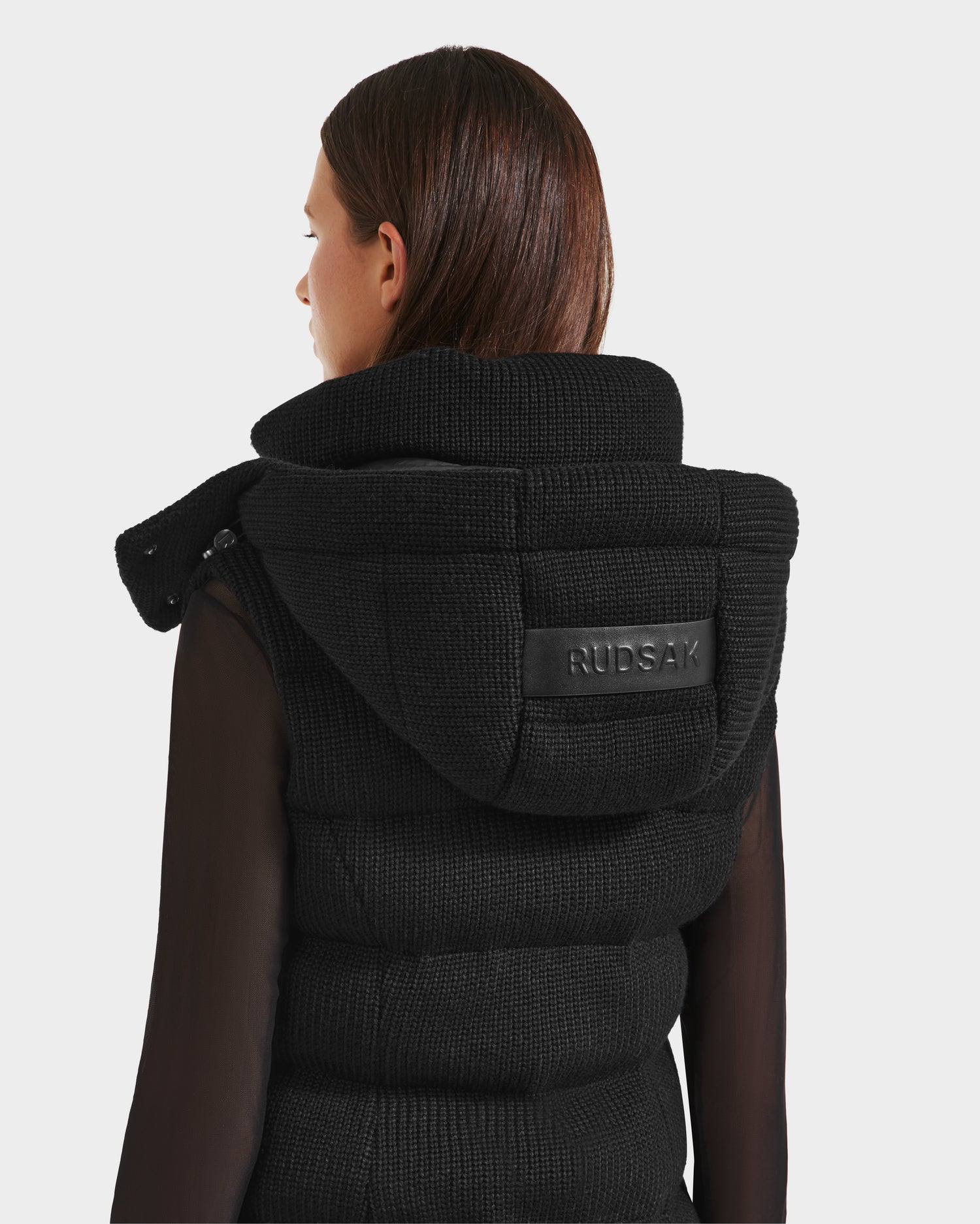 A person with straight brown hair faces away from the camera, wearing the Rudsak CASSIDY - 8125622 Black women’s fitted down vest with a removable hood and leather label, layered over a sheer black long-sleeve top, against a plain backdrop.